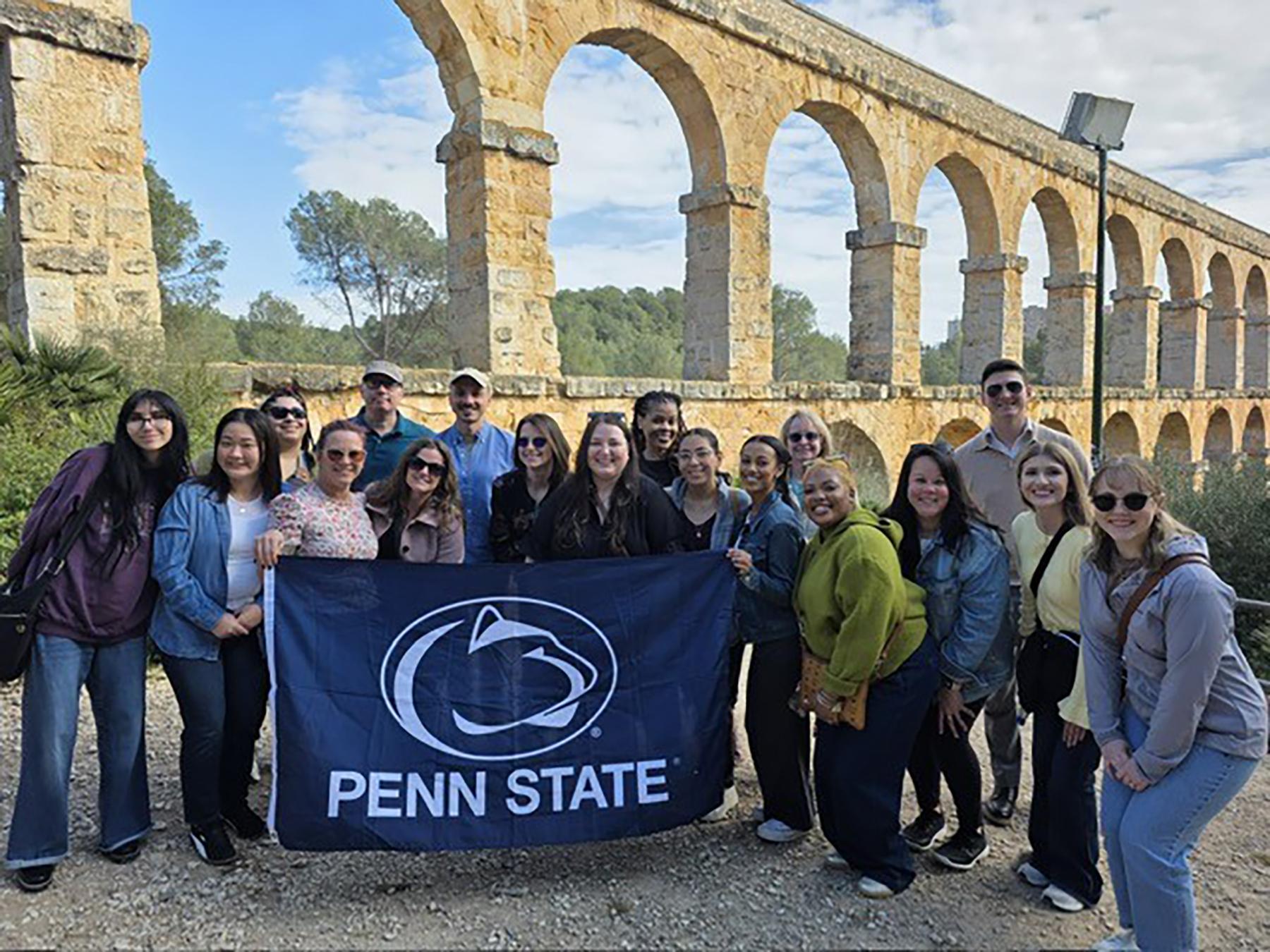 A group of people holding a navy Penn State flag stand in front of a rock bridge.