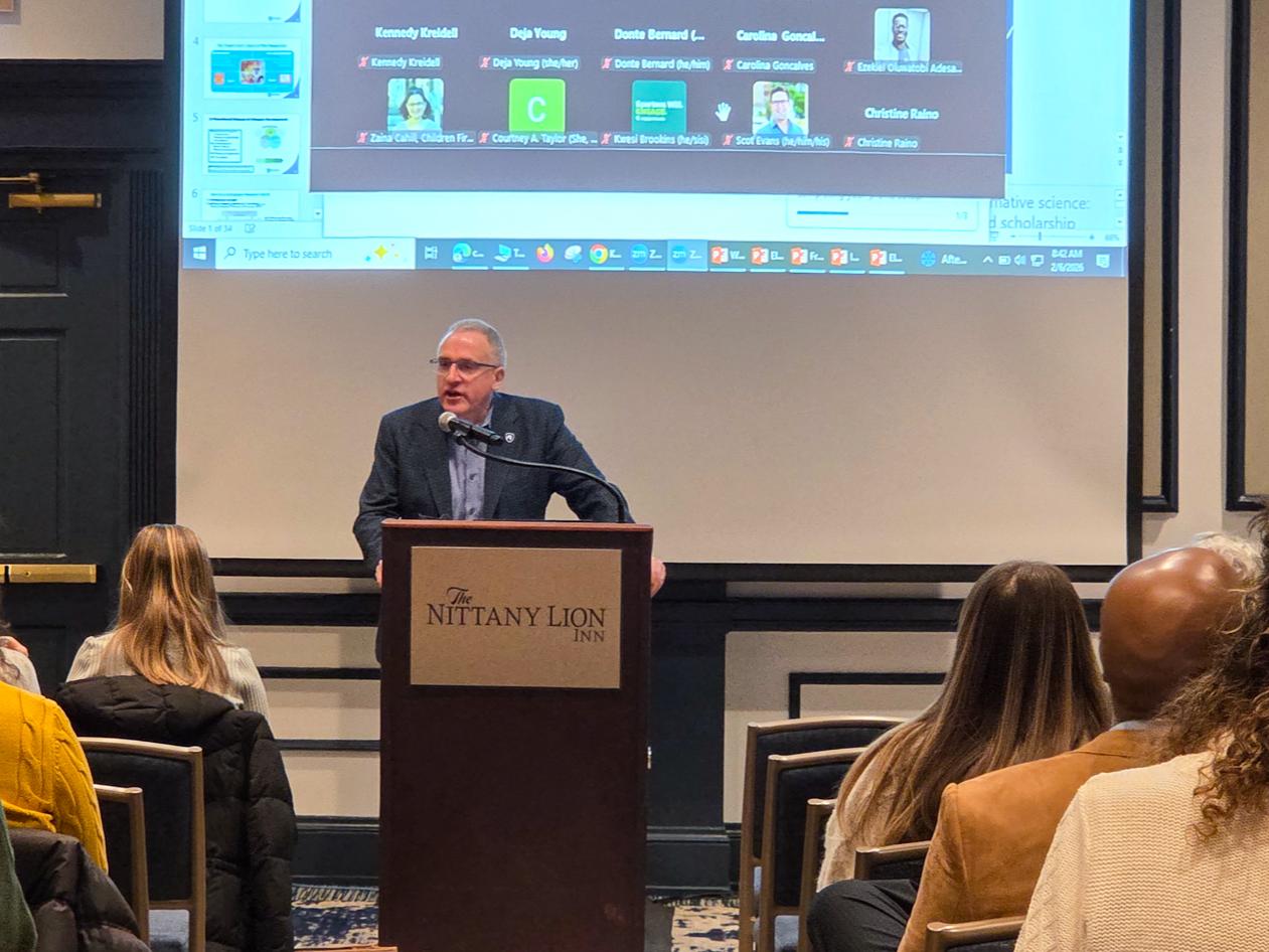 A presenter stands behind a podium at the front of a conference room while attendees sit in rows facing forward. A large projection screen shows a virtual meeting interface with numerous participant icons arranged in a grid format. Slide thumbnails appear along the left side of the screen, suggesting the presenter is sharing a computer desktop. The room has dark-paneled doors and trim, with neutral-colored walls.