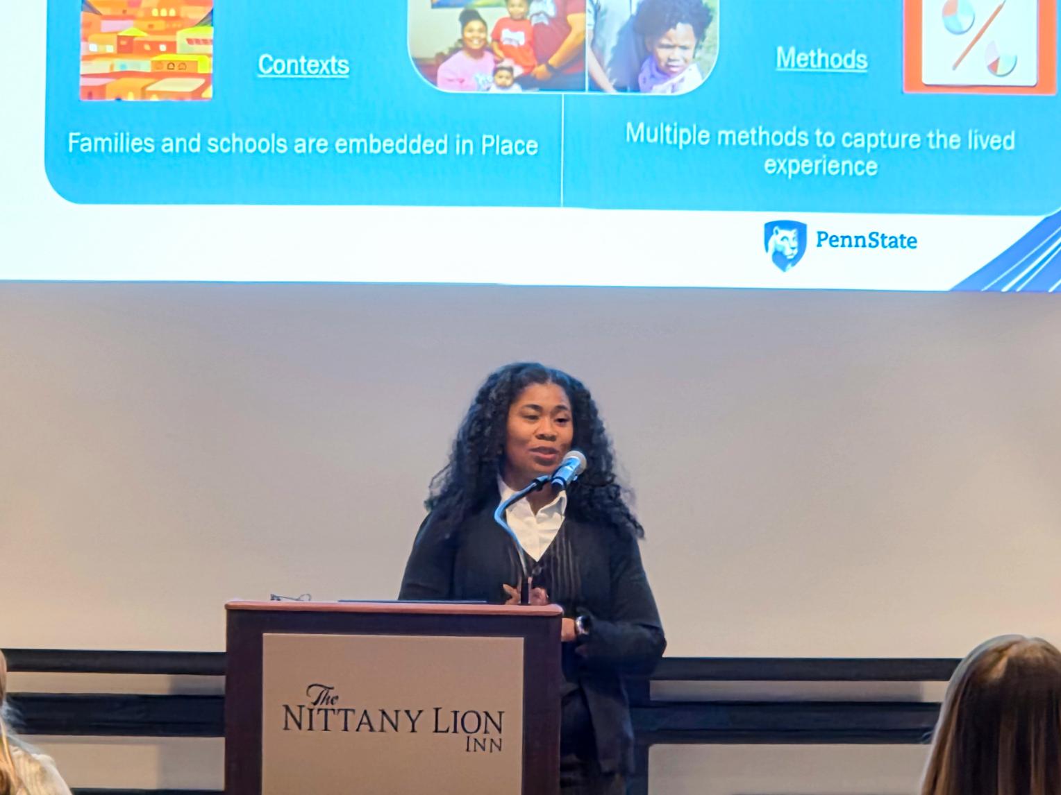 A presenter stands behind the podium giving a presentation about family and school research contexts. The slide behind the presenter features colorful graphic icons including books, family photos, and symbols representing research methods. The slide divides content into labeled sections such as “Contexts” and “Methods.”