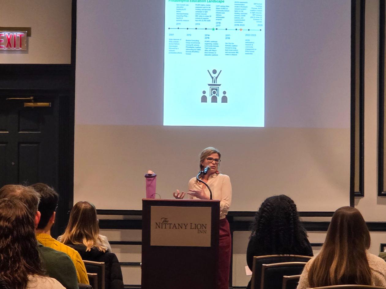 A speaker stands at a podium presenting a slide with a family photo and text about community-engaged science, while attendees sit facing the screen.