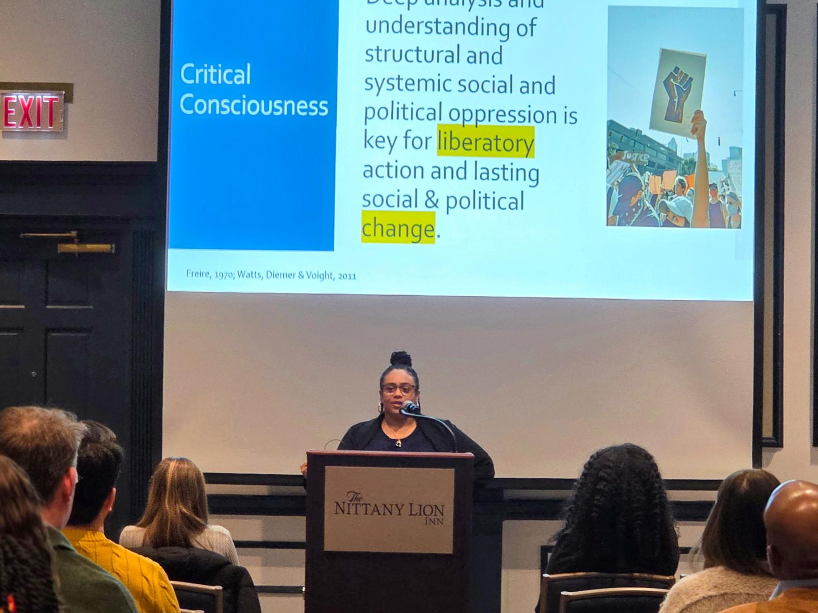 A speaker stands at a podium presenting a slide with a family photo and text about community-engaged science, while attendees sit facing the screen.