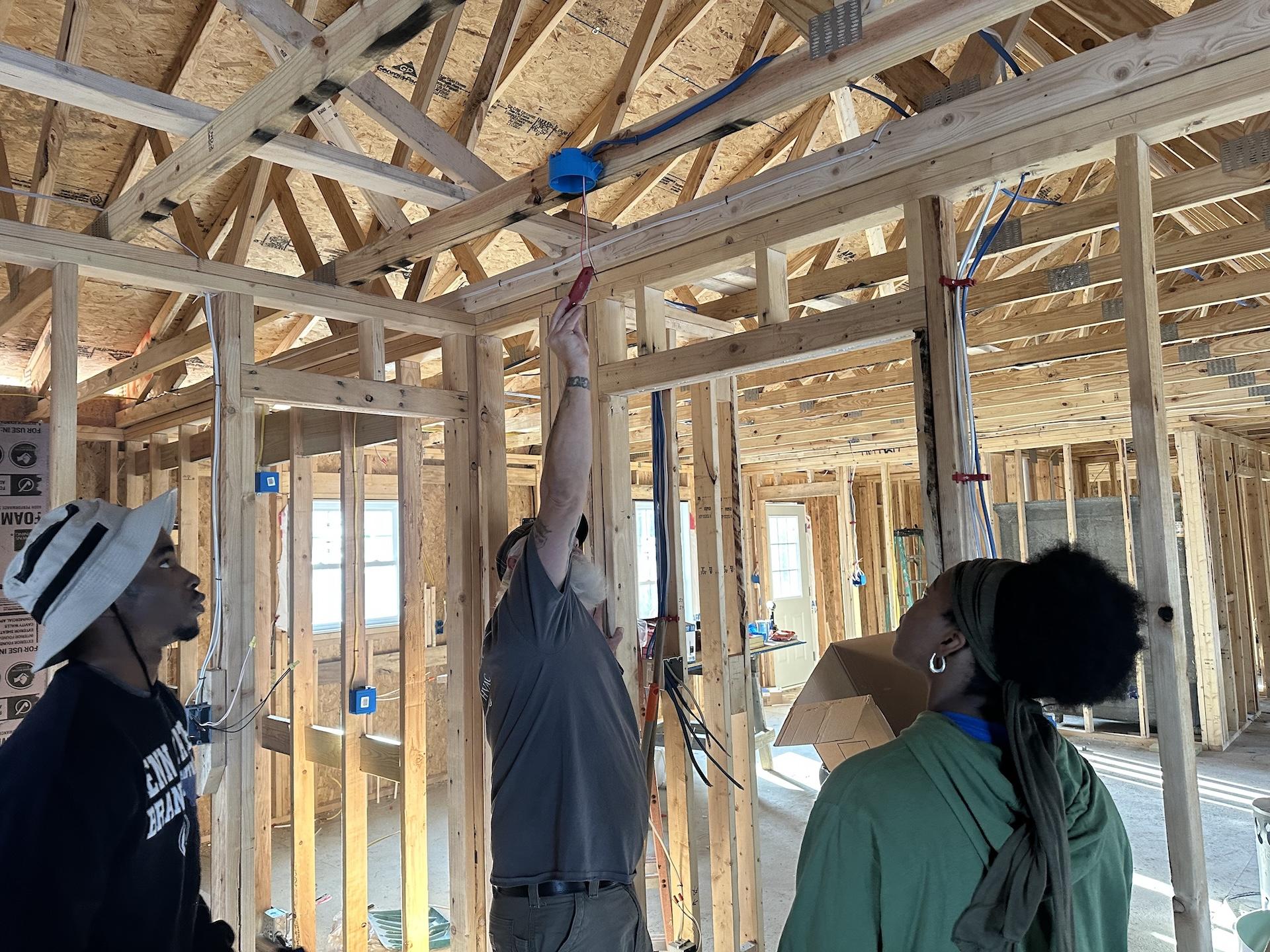 two students watching a Habitat for Humanity volunteer drilling wood into place in the ceiling