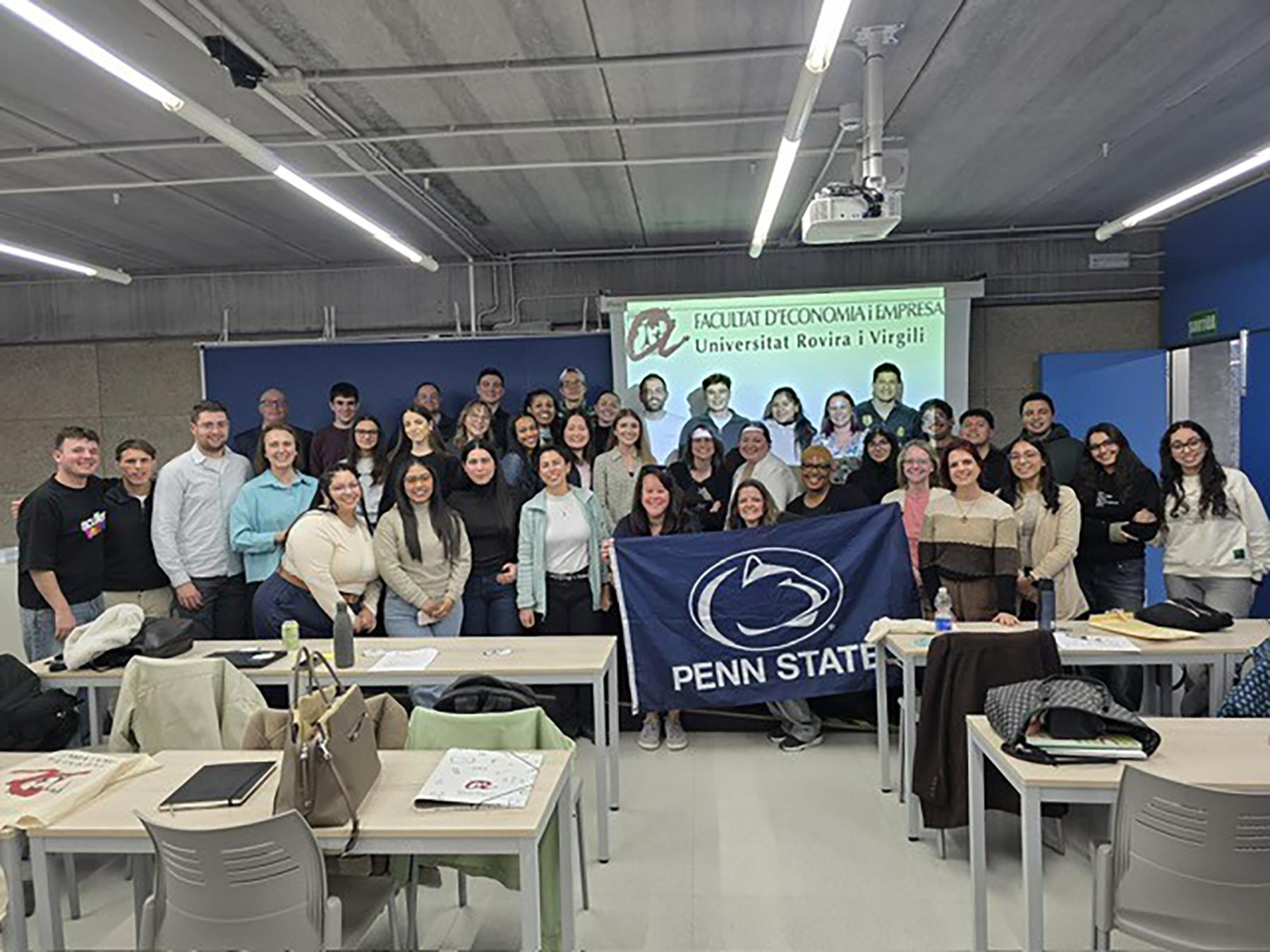A group of people holding a navy Penn State flag in a classroom.