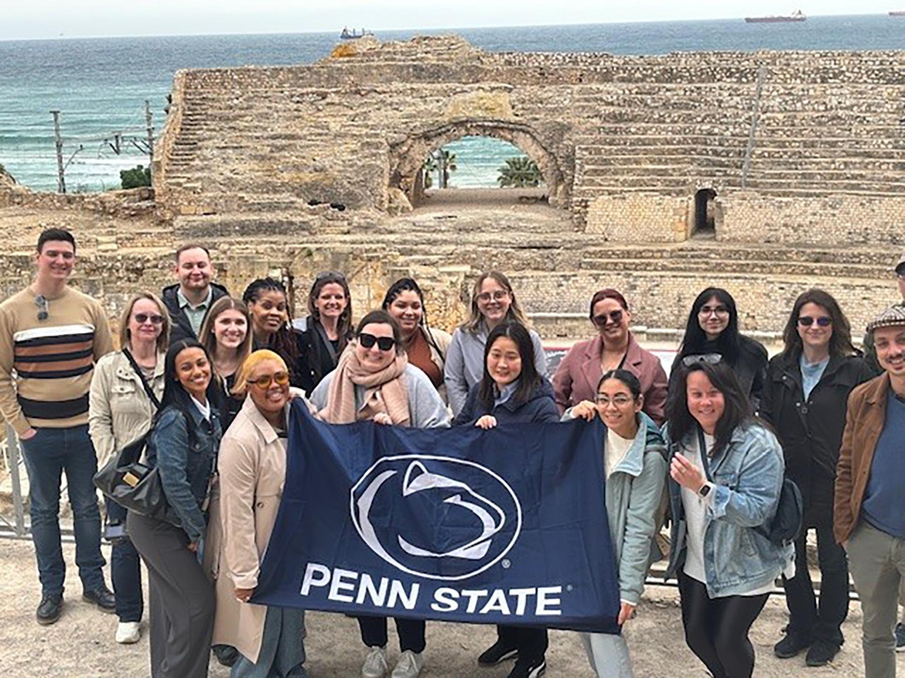 A group of people holding a navy Penn State flag pose in front of a stone historical site.