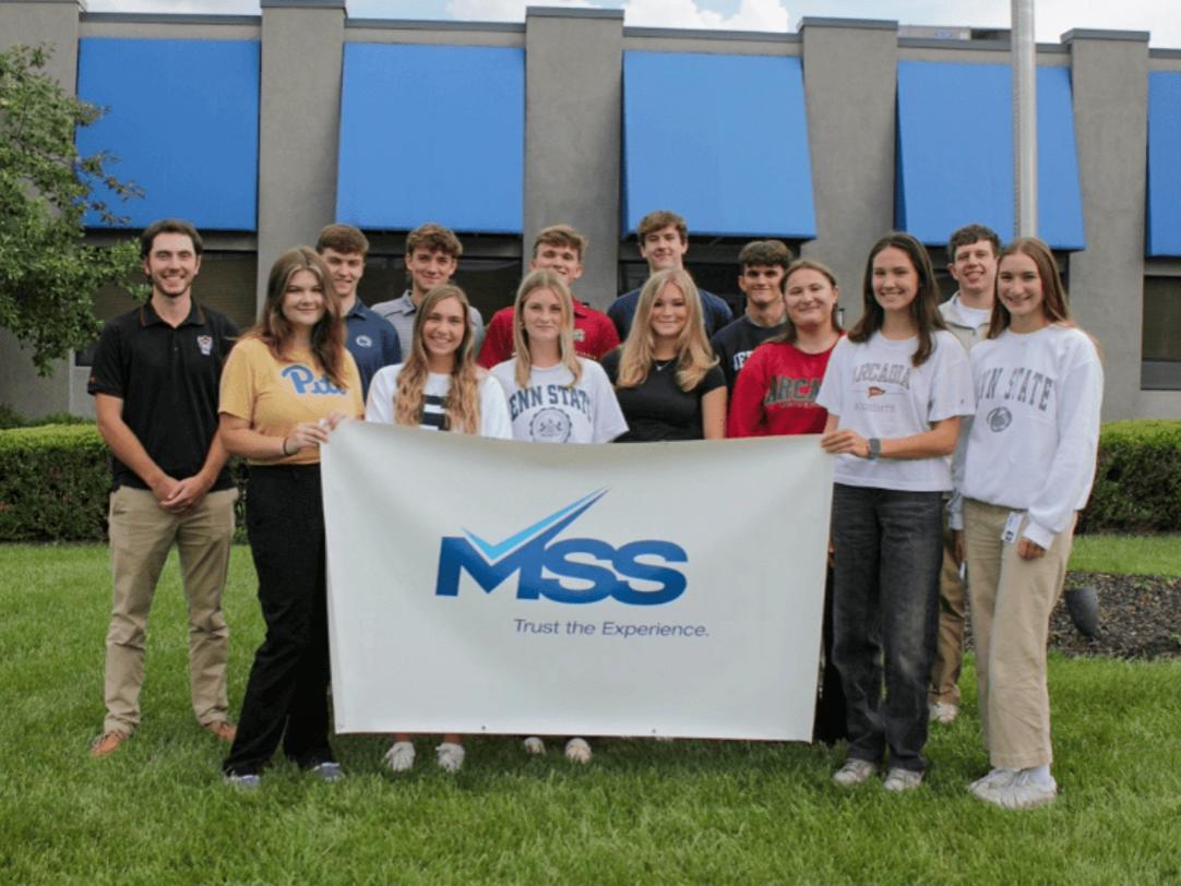 Jessica Amatrudo wears a white S sweatshirt while standing with a group of interns behind a white banner that reads “MSS Trust the Experience.” 