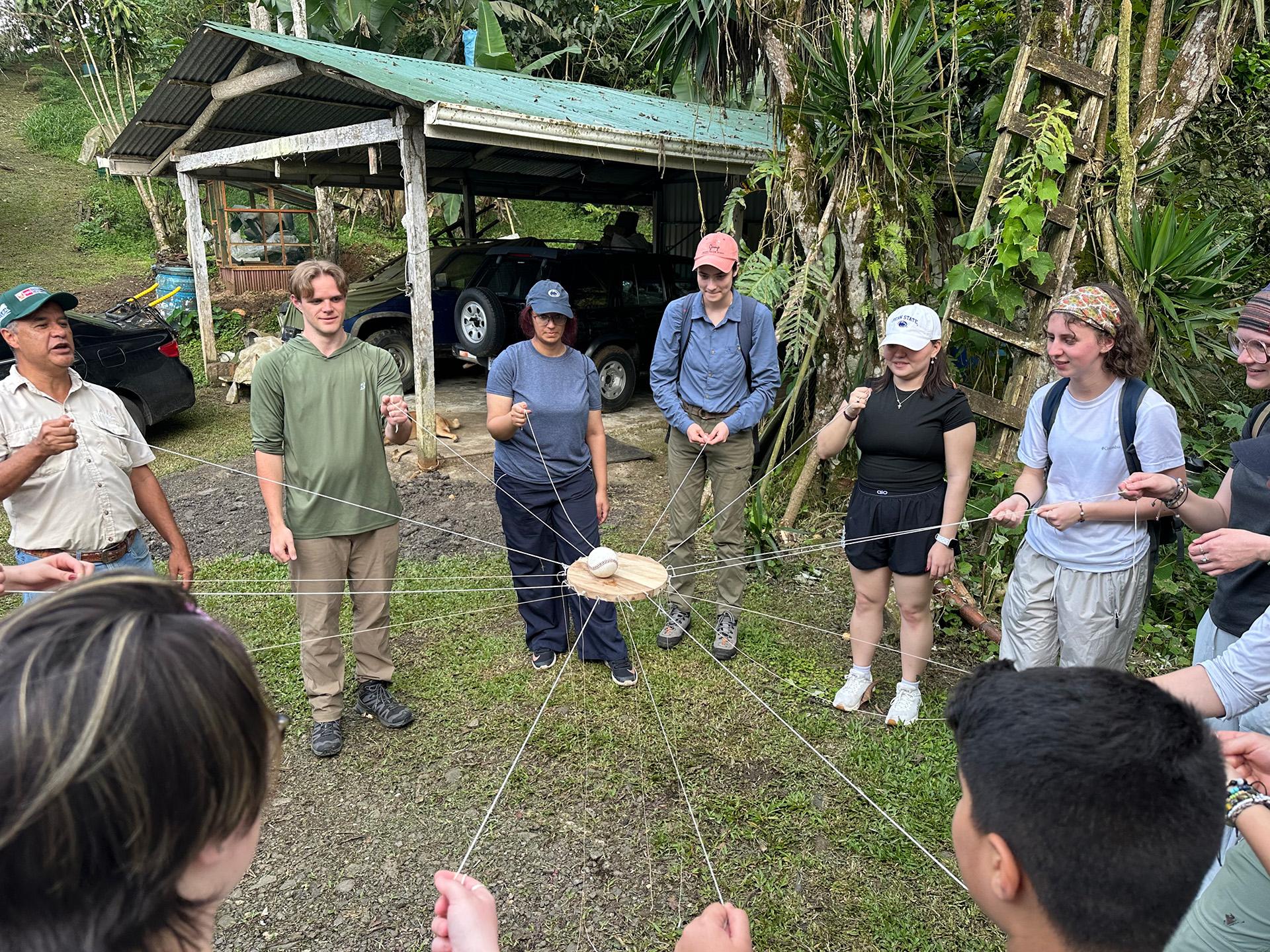 People standing in a circle holding strings, the strings are attached to a piece of wood being held up in the center of the circle