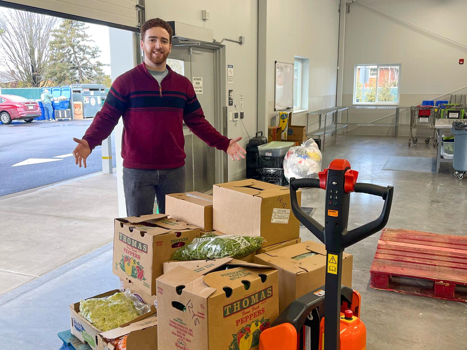 Emmanuel Kraft stands arms wide behind stacks of boxes filled with food.