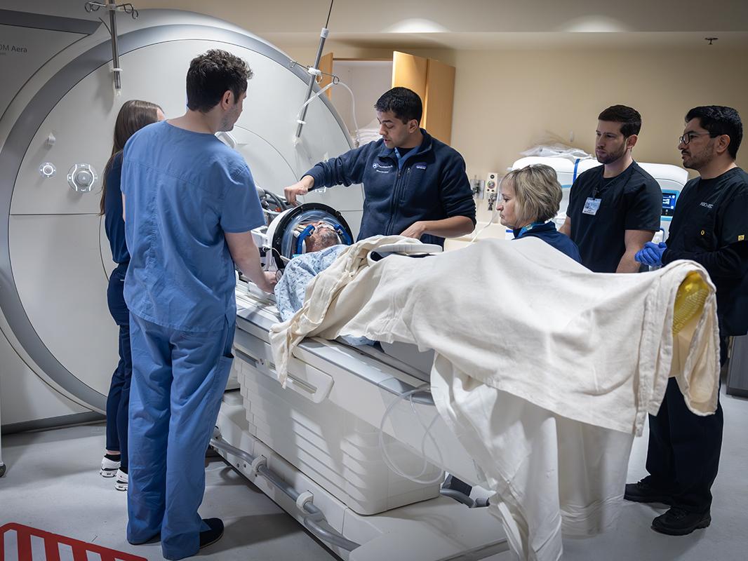 A group of five MRI technologists and one physician stand next to a male patient who is about to go into an MRI machine. The physician, center, points to the patient.