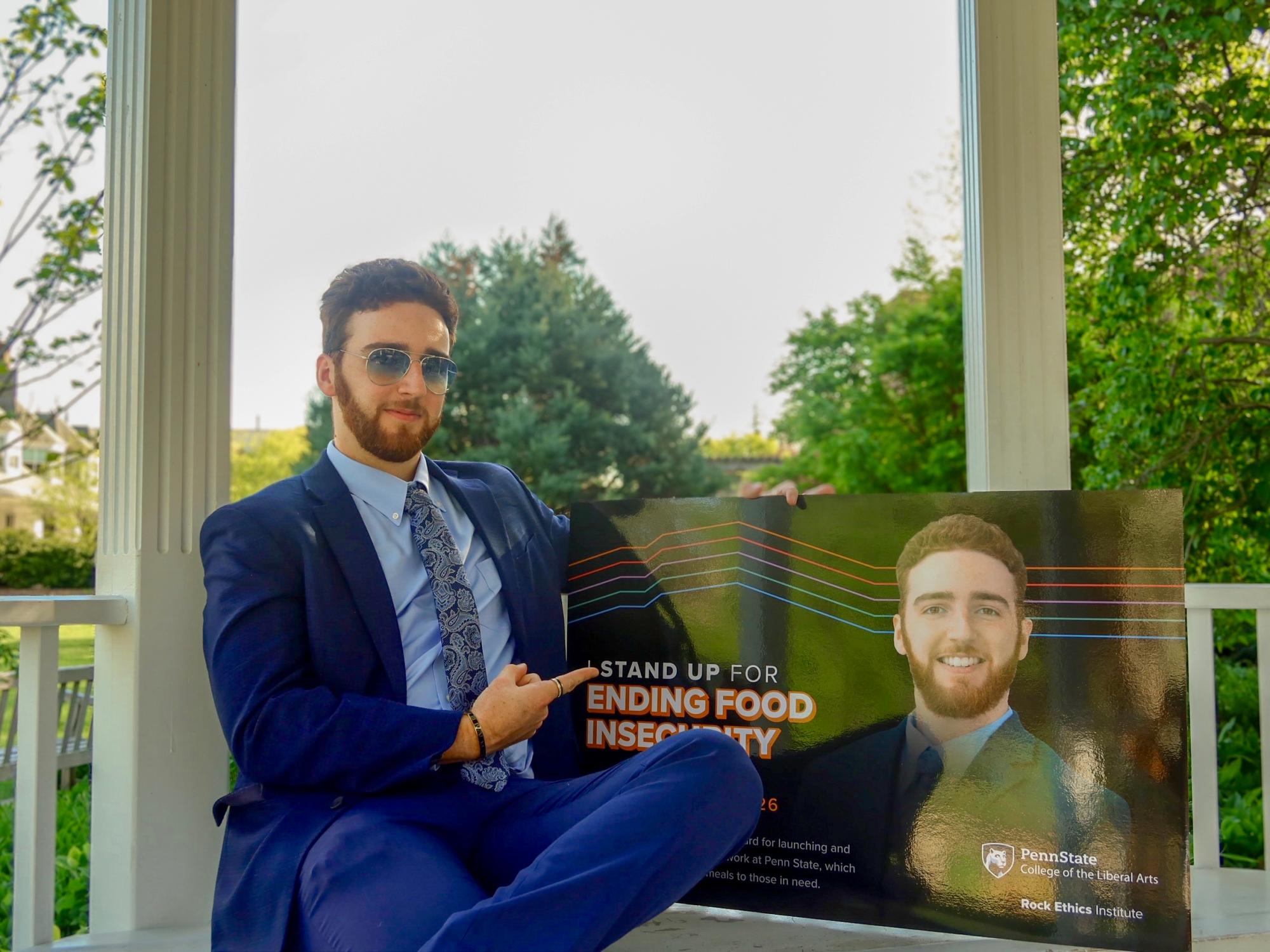 Emmanuel Kraft, wearing a blue suit and sunglasses, sits outside on a gazebo bench while pointing to a poster with his face on it.