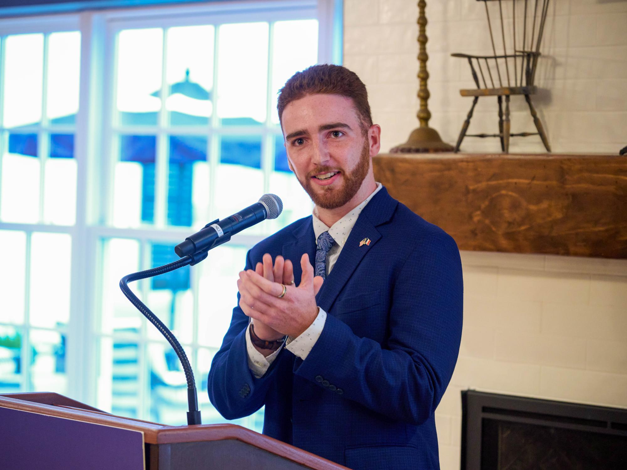 Emmanuel Kraft speaks into a microphone at a podium during a campus event, wearing a blue suit and addressing an audience.