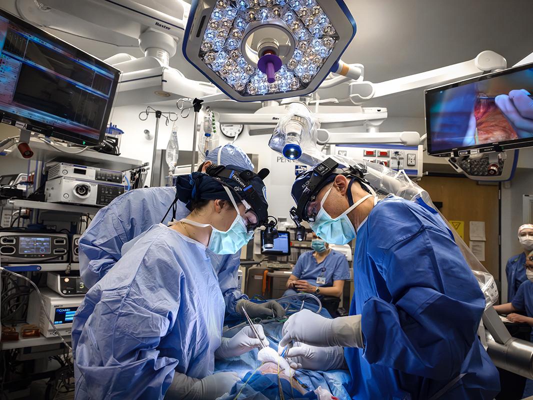A surgeon, surgical resident and surgical assistant wearing surgical gowns and masks perform surgery on a patient at Penn State Health Milton S. Hershey Medical Center. Two monitors are on the left and right side of the room, and a light is above them. 