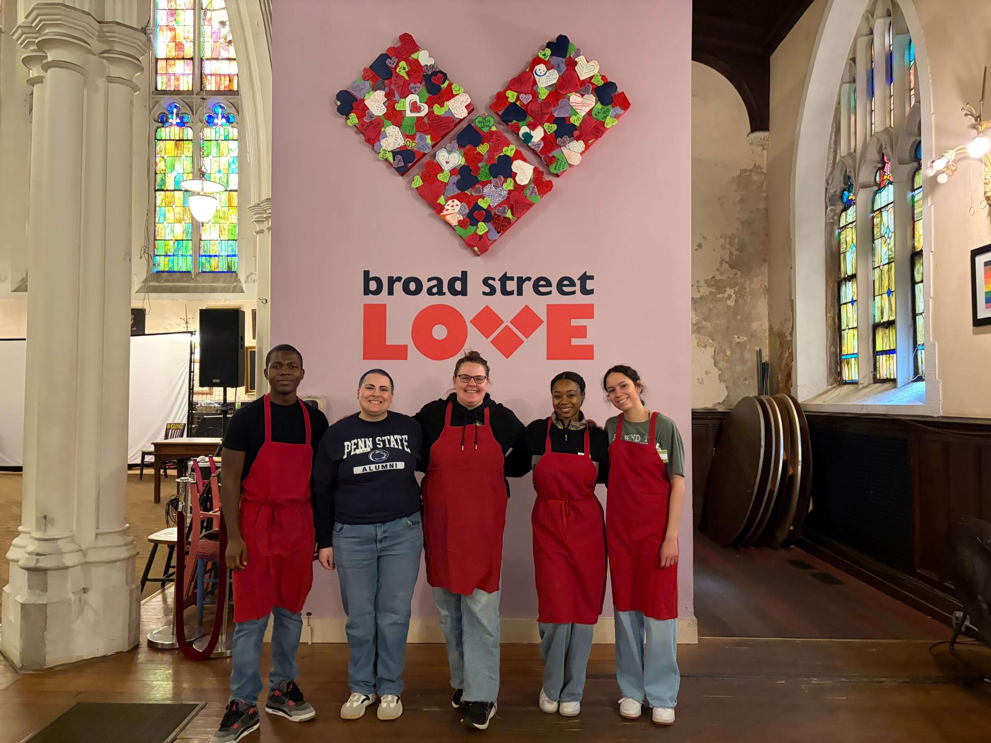Five people standing in front of a white wall with a heart and the words Broad Street Love printed on it.