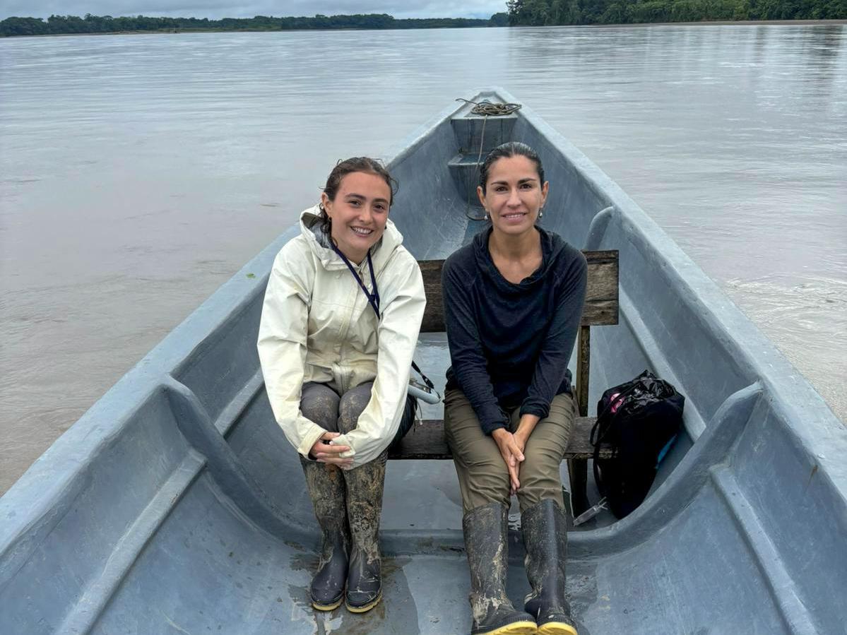 Two women sit on a wooden bench in a gray boat on a wide river under a cloudy sky. They wear boots and are smiling.