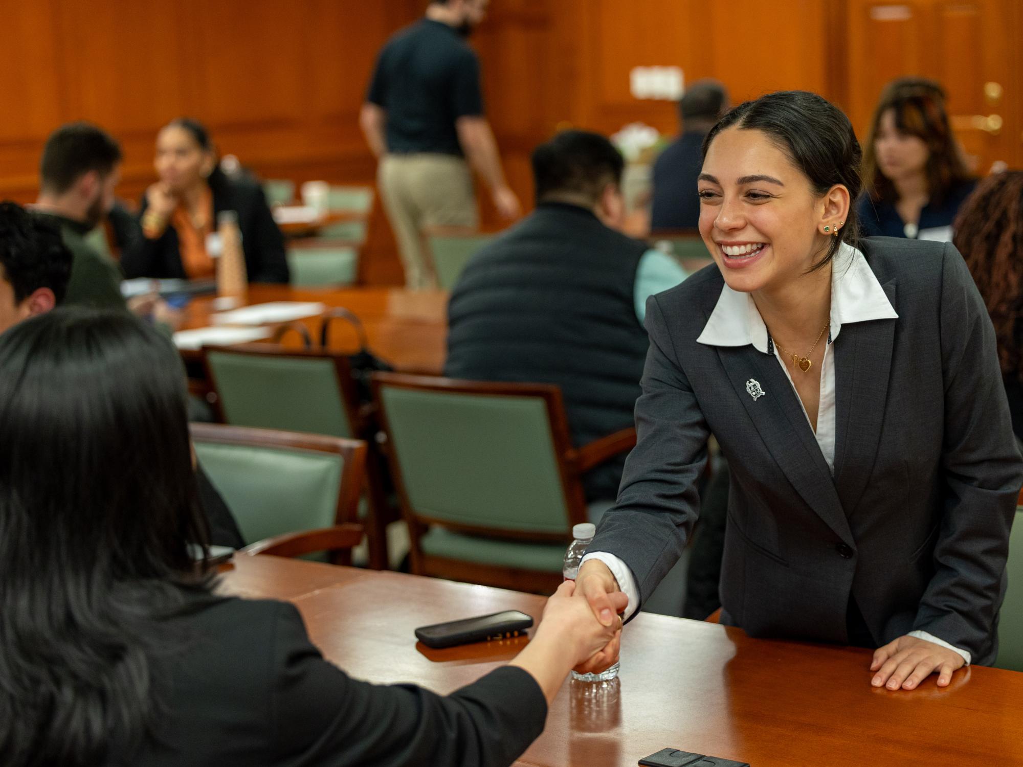 Two smiling women in business dress shake hands over a wooden table. 