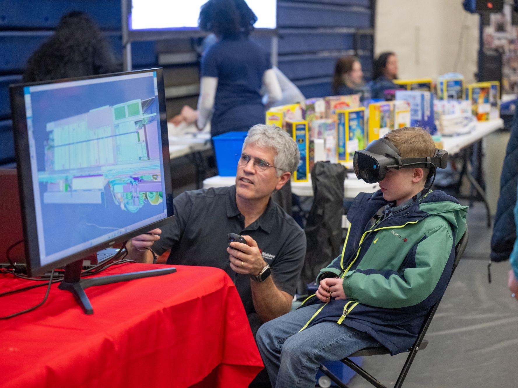 A man helps a boy in a virtual-reality headset navigate a locomotive simulation.