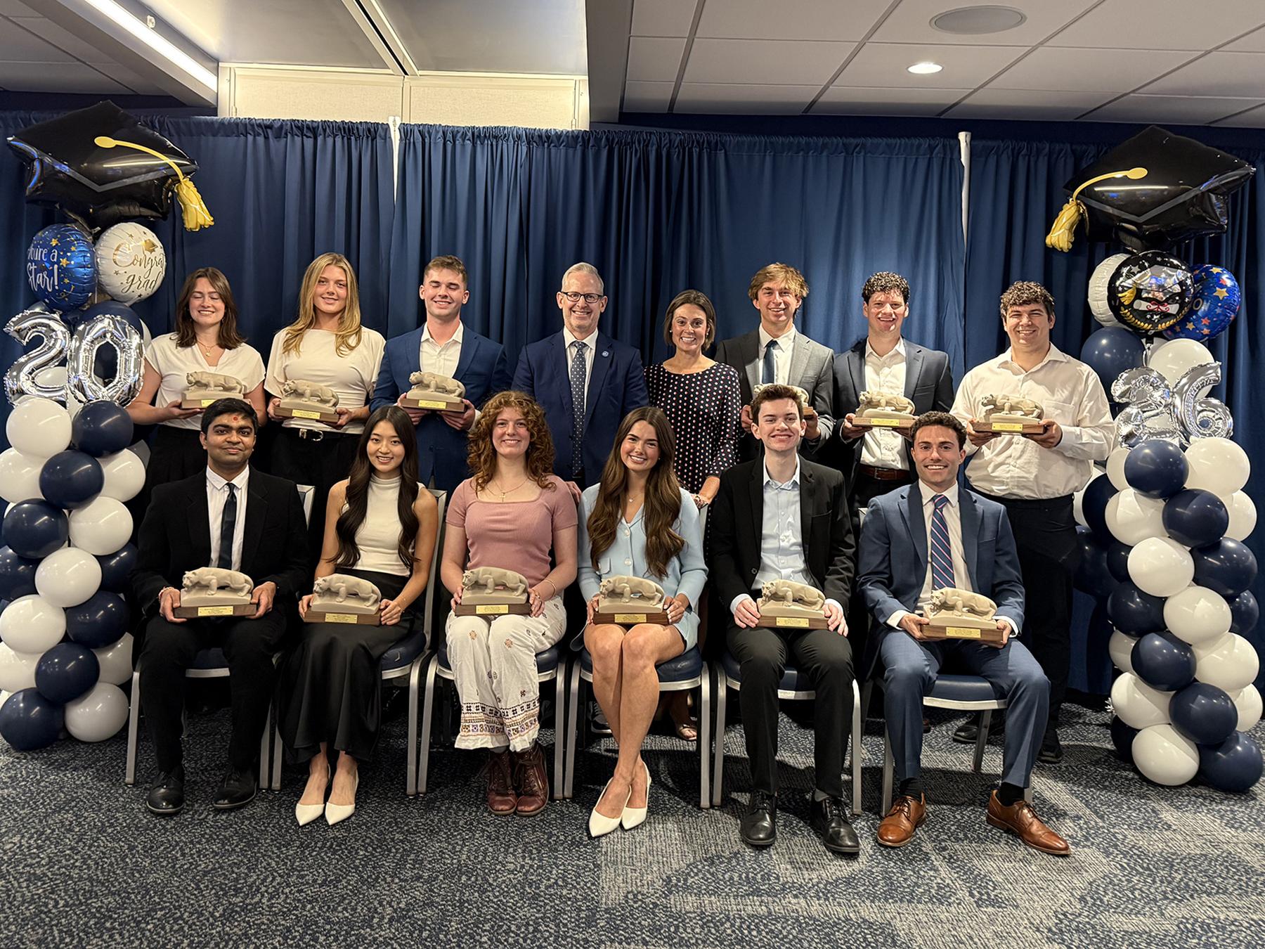A photo of six male and six female students seated and arrayed around Dean Corey Phelps and Associate Dean for Undergraduate Education Jennier Eury seated and standing with balloons on either side of the group.