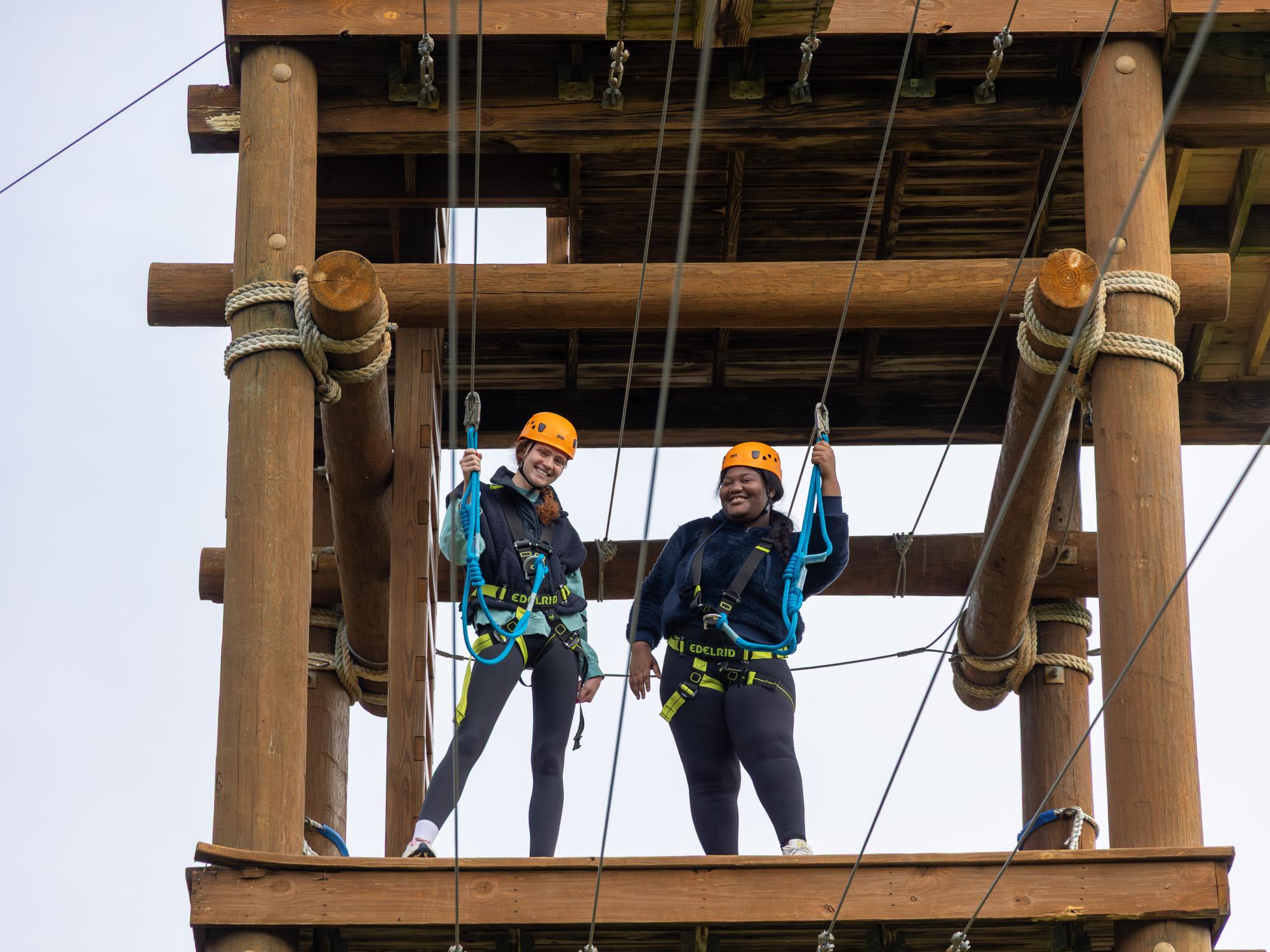 Two students stand on a platform while strapped into harnesses on an aerial ropes course.
