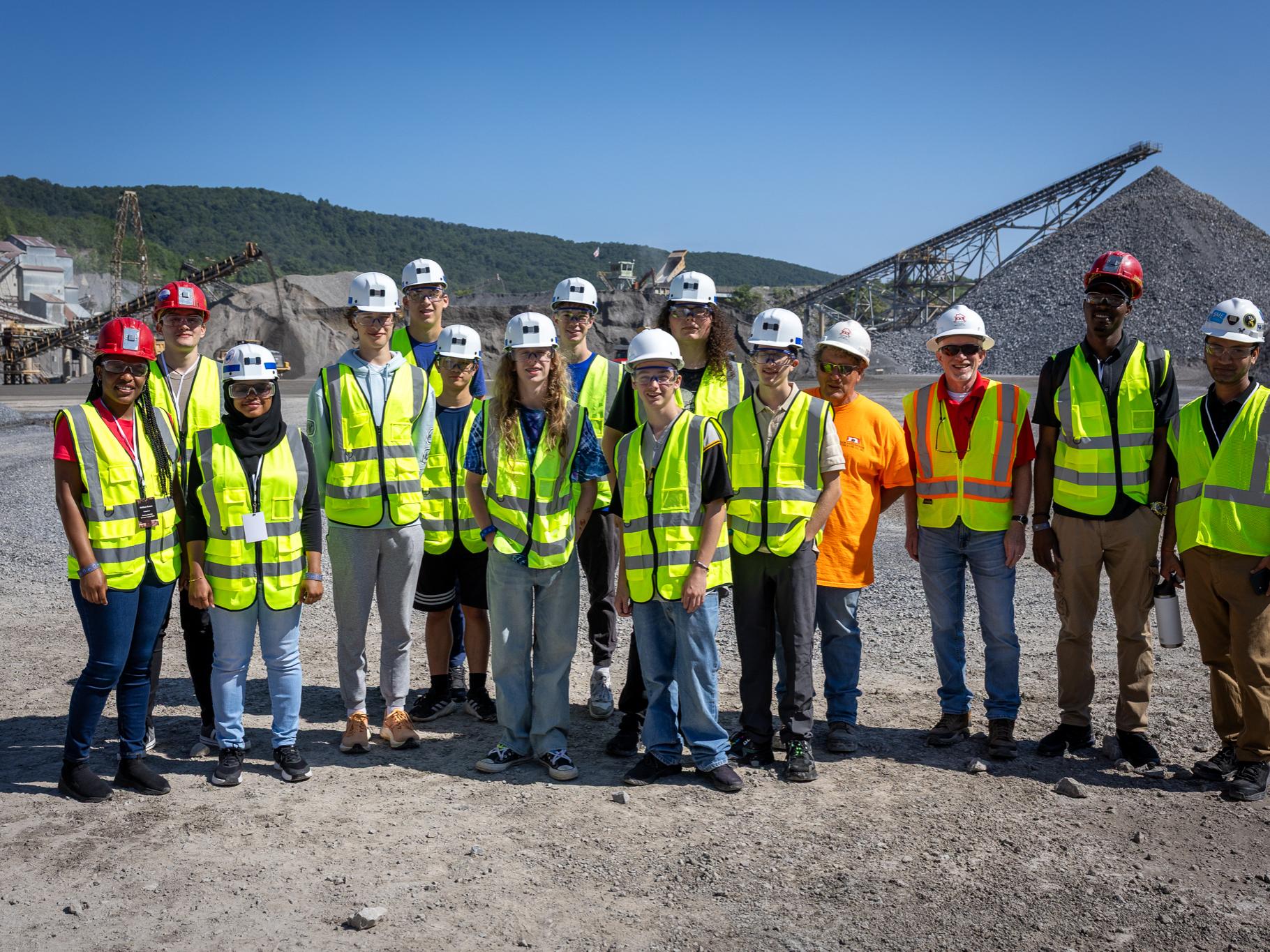 A large group of students wearing hardhats and safety vests pose for a photo at a rock quarry. 