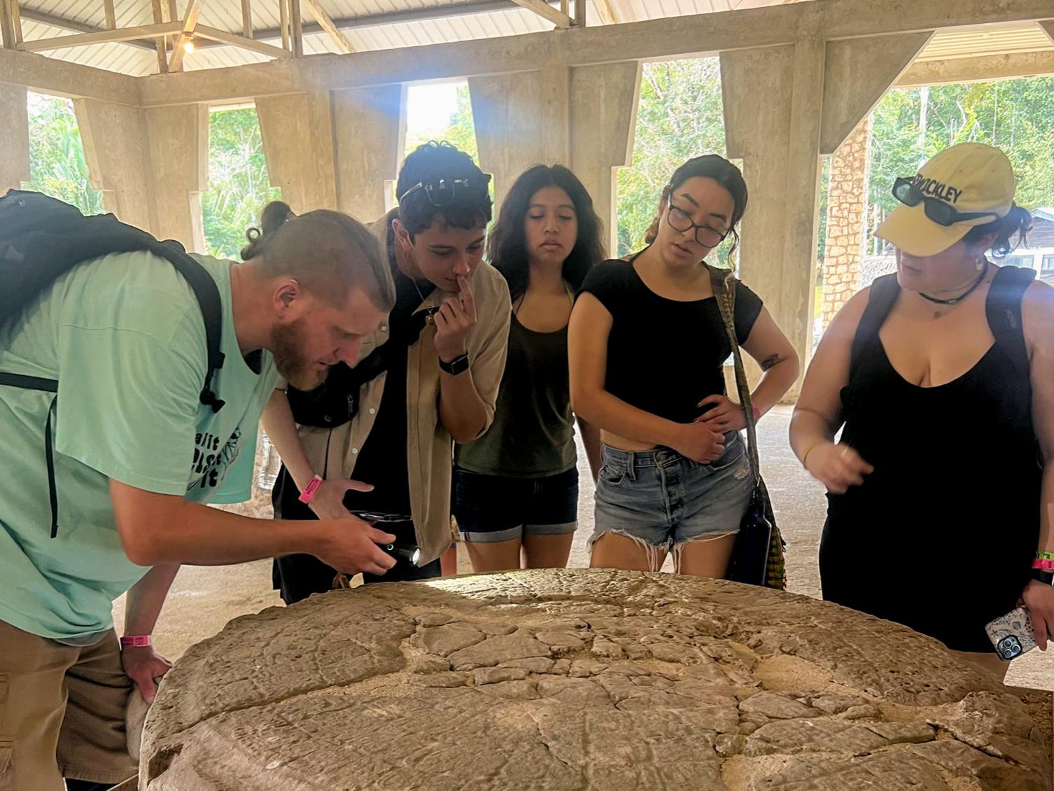 Several people stand around a large circular carved stone. They lean in to study the weathered carvings on its surface.