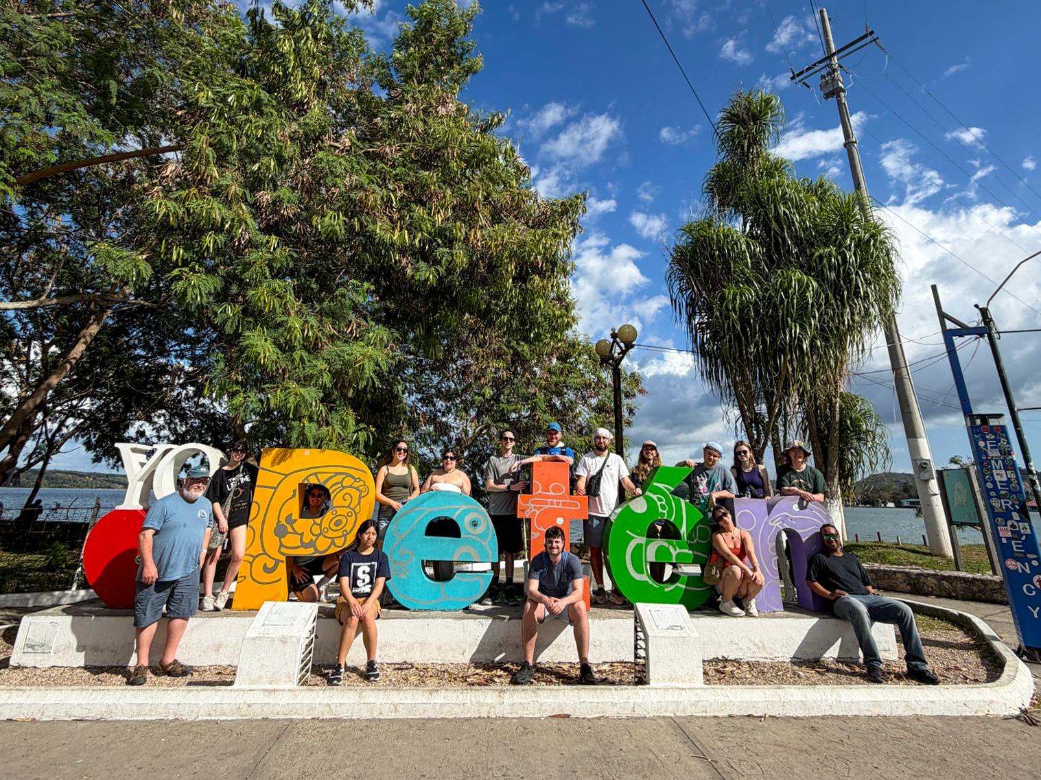 A group of people pose in front of a colorful “Yo Heart Petén” sign near a lakeside walkway. Each letter is decorated with bright patterns, and trees and water fill the background under a partly cloudy sky.