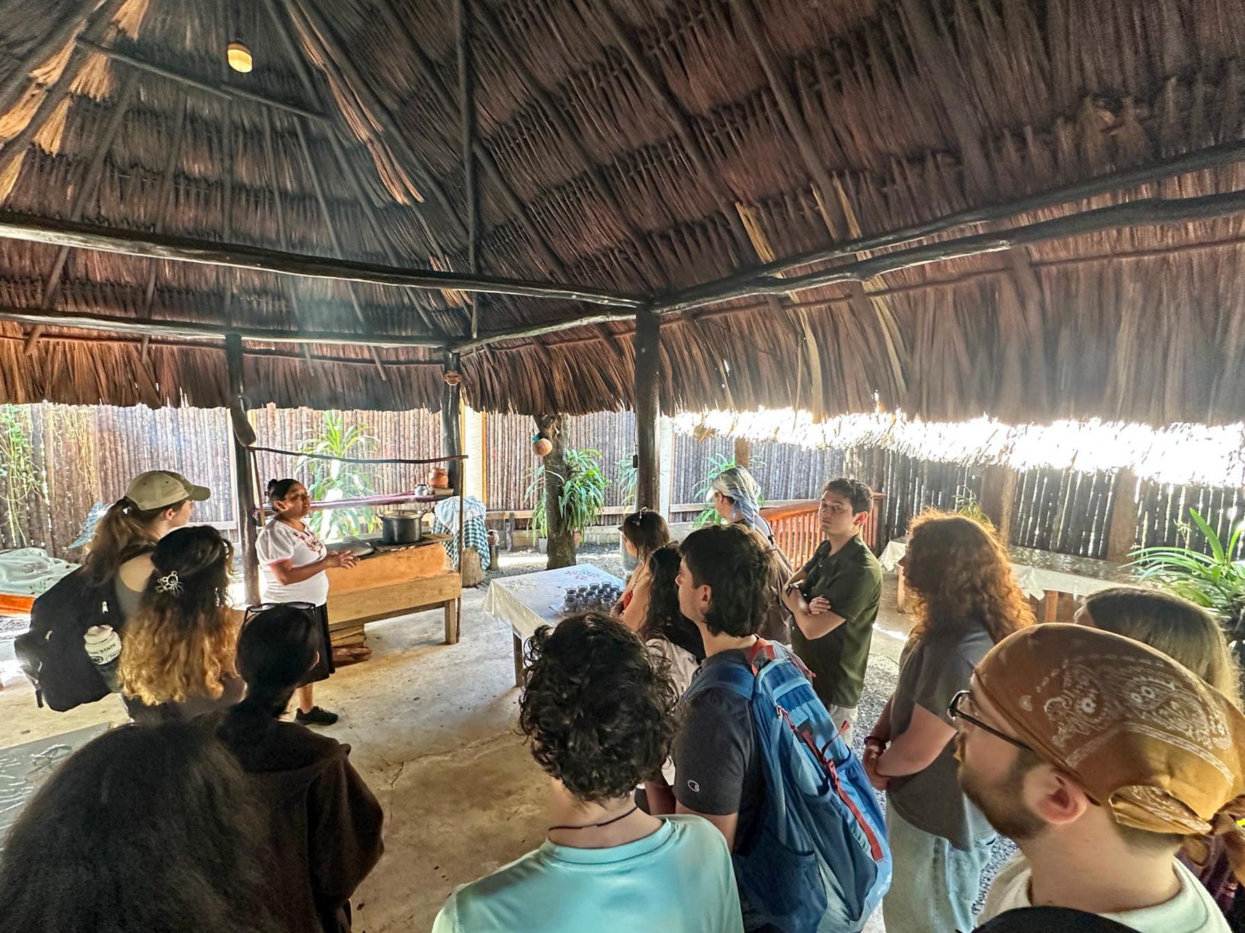 A group of people gathers inside a thatched-roof structure watching a woman lead a cooking or cultural demonstration at a table. Natural light filters through the open sides of the hut, and the audience stands closely, observing.