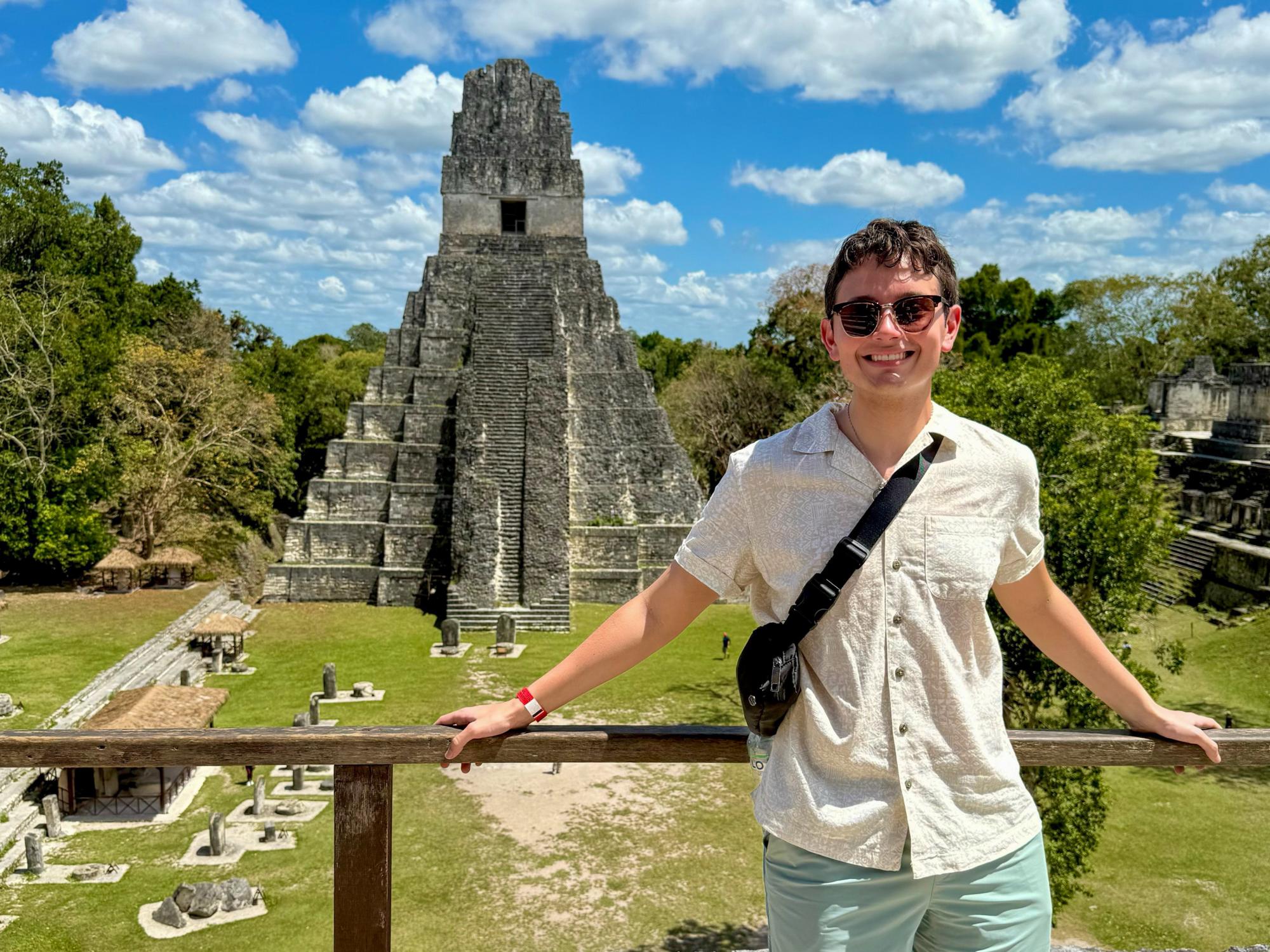 A person wearing sunglasses, a light shirt, mint green shorts, and a crossbody bag stands on a wooden platform overlooking a Tikal pyramid. The sky is blue with scattered clouds.