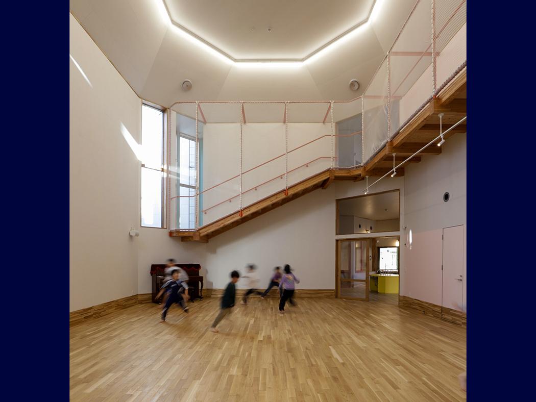 The inside of a well-lit spacious room with wood flooring, a stairwell and what appears to be people in motion.
