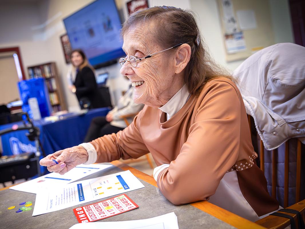 A woman smiles as she holds a bingo chip. Worksheets and a bingo card are in front of her on a table. She is wearing glasses, a turtleneck top and a long-sleeved top.
