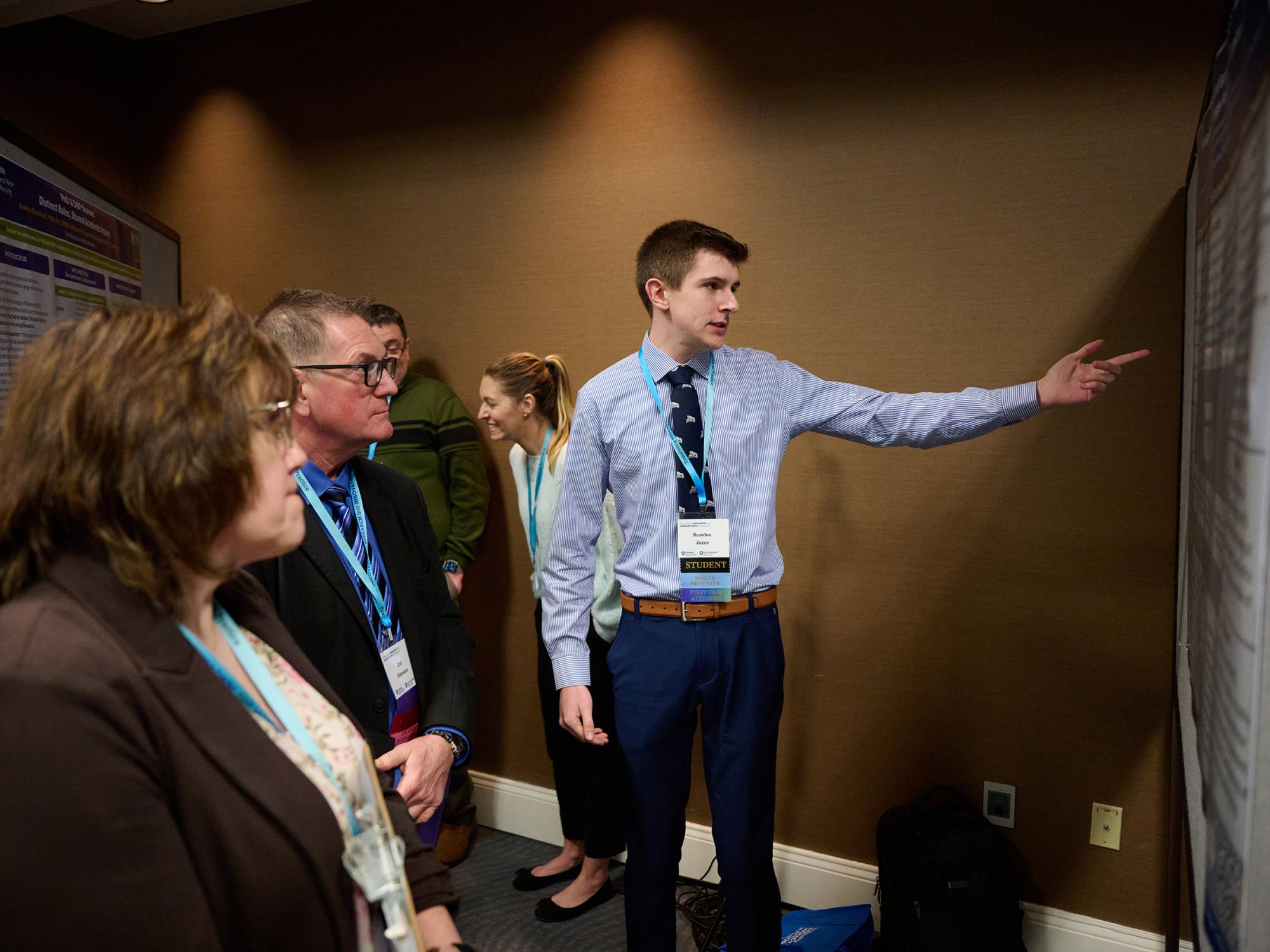 Braedon Joyce, at right, gestures to information on his research project display to three attendees at the annual symposium