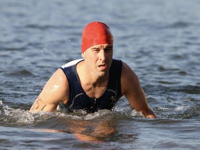 A man swims in the ocean during a triathlon. He is wearing a bathing cap and sleeveless wetsuit.
