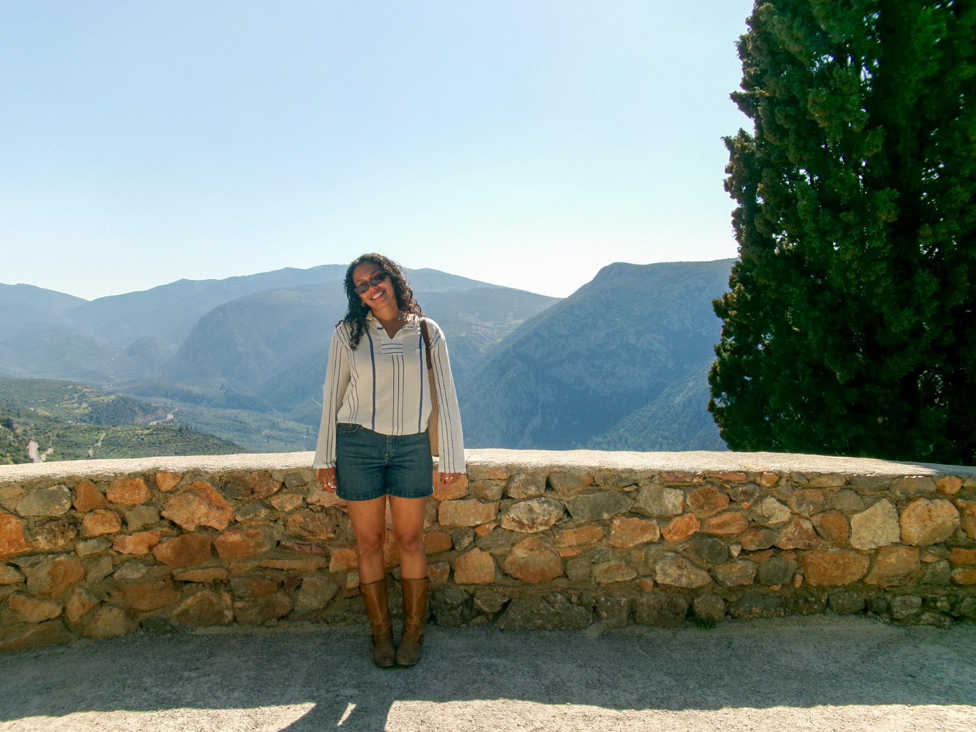 Jasmin Tanner wears a white blouse, jean shorts, and brown boots with mountains in the background.