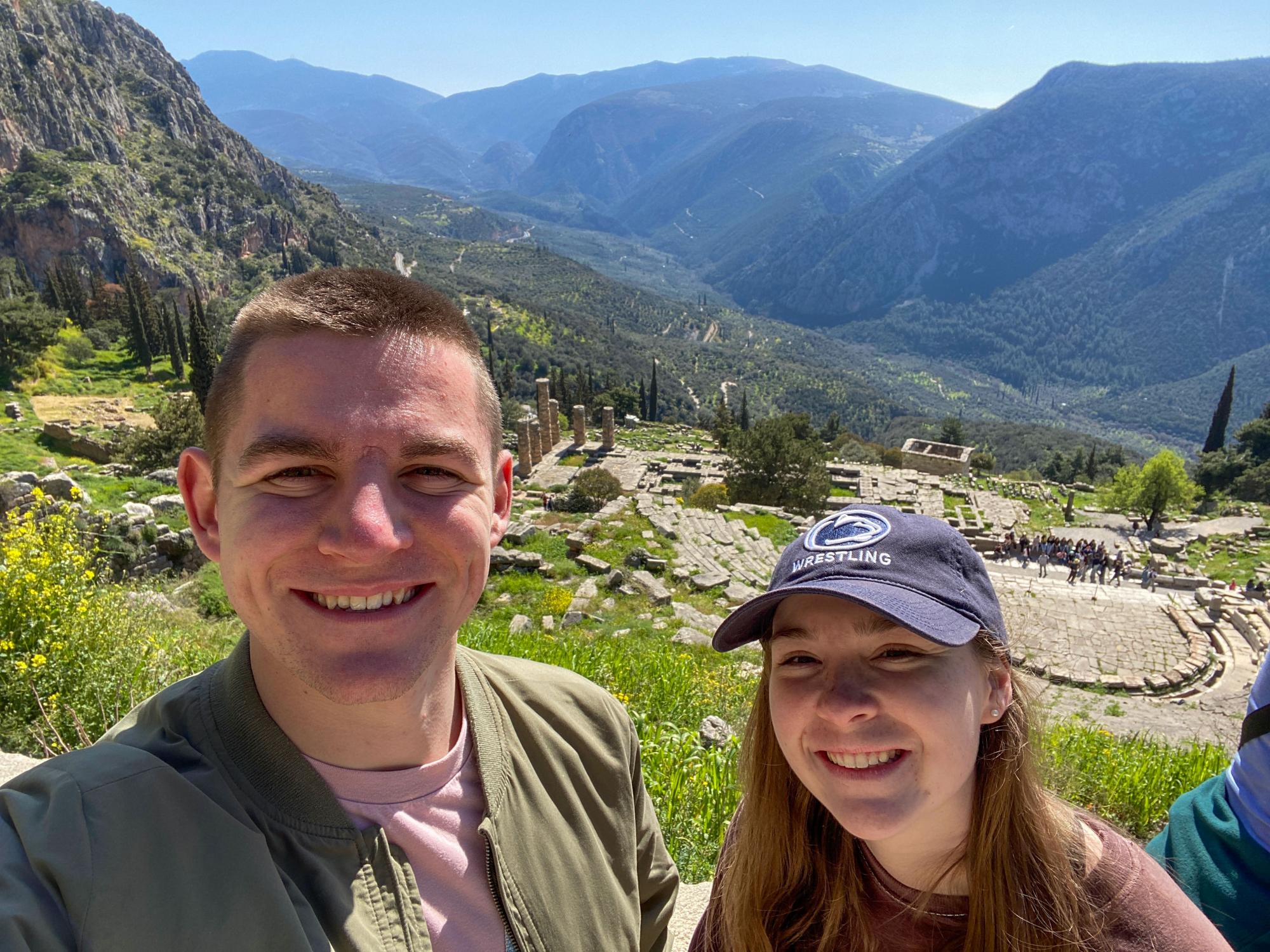 Luciano Ragonese and Ireland McDyre stand in front of the Sanctuary of Apollo. Luciano is wearing a green jacket and pink shirt. Ireland wears a Penn State wrestling hat.