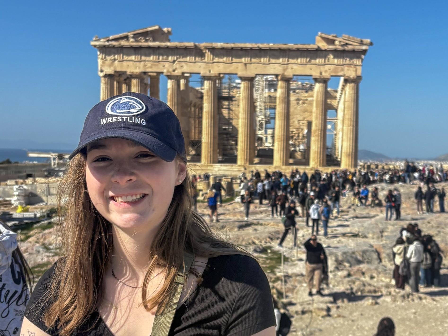 Ireland McDyre wears a black shirt, Penn State hat, and green fanny pack with the Parthenon on the Acropolis in the background.