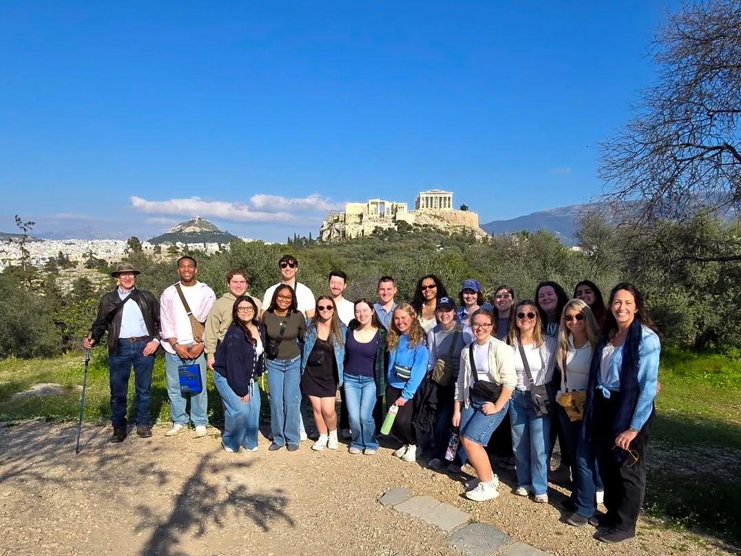 : The group stands by the rocks in front of Pnyx, Areopagus, with Professor Munn holding a walking stick on the left and Professor Smagh on the right of the group.