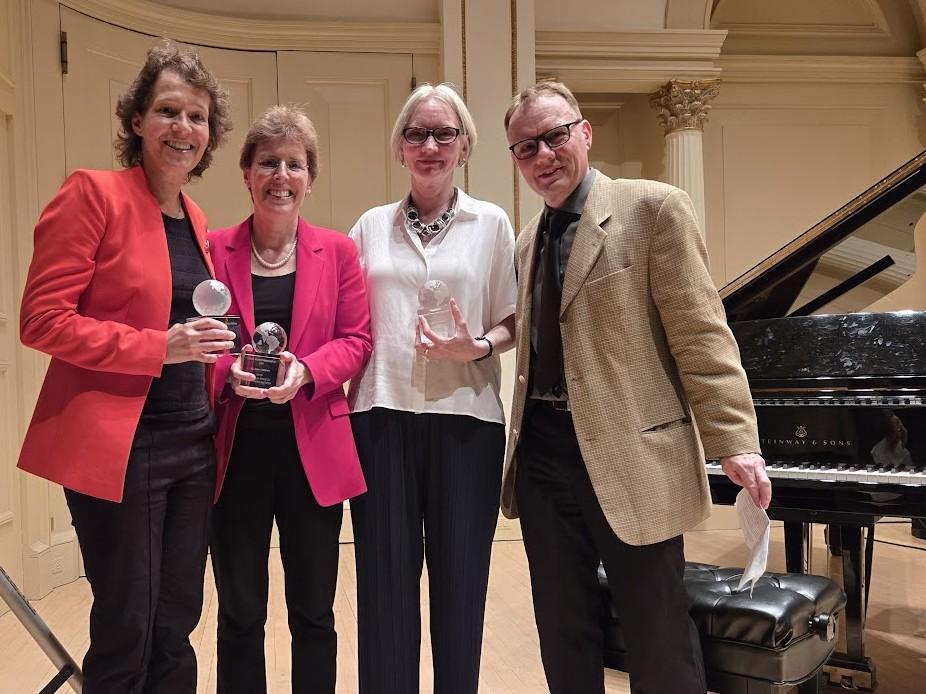Four individuals smile together in Carnegie Hall, three hold awards