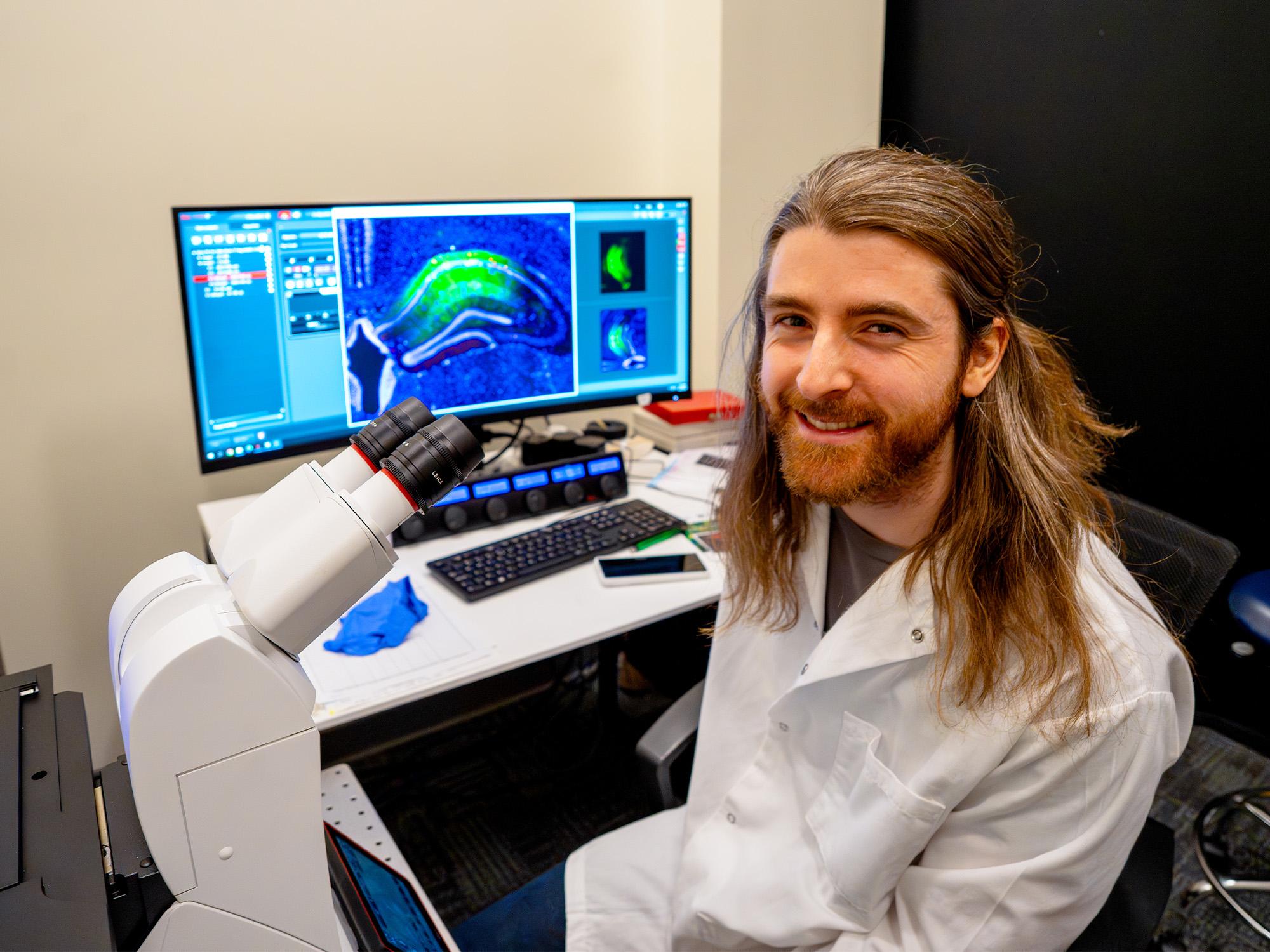 person sits next to microscope and computer monitor with brain image displayed