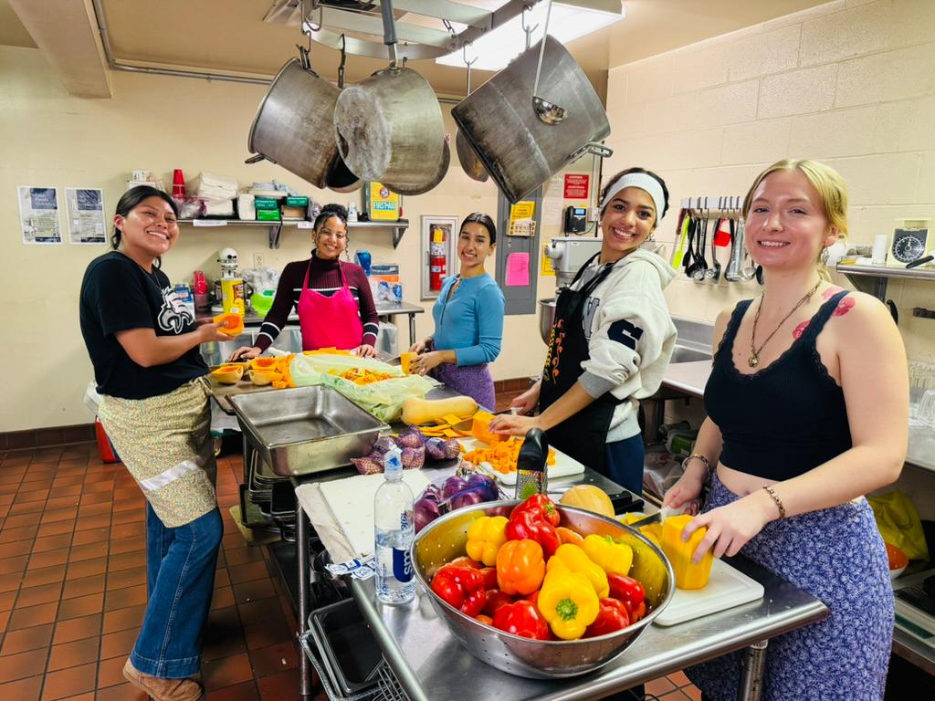 : Laenee McCoy prepares food in the kitchen with fellow IPSA group members. Laenee is wearing a black tank top and purple pants.