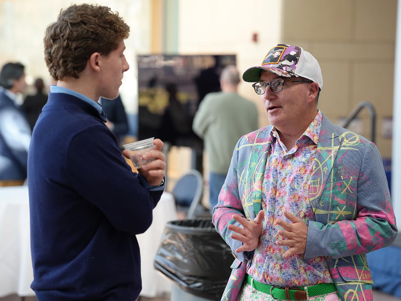 Dan Ives, wearing a hat and mulitcolored suit, speaks with a student in the Business Building Atrium.