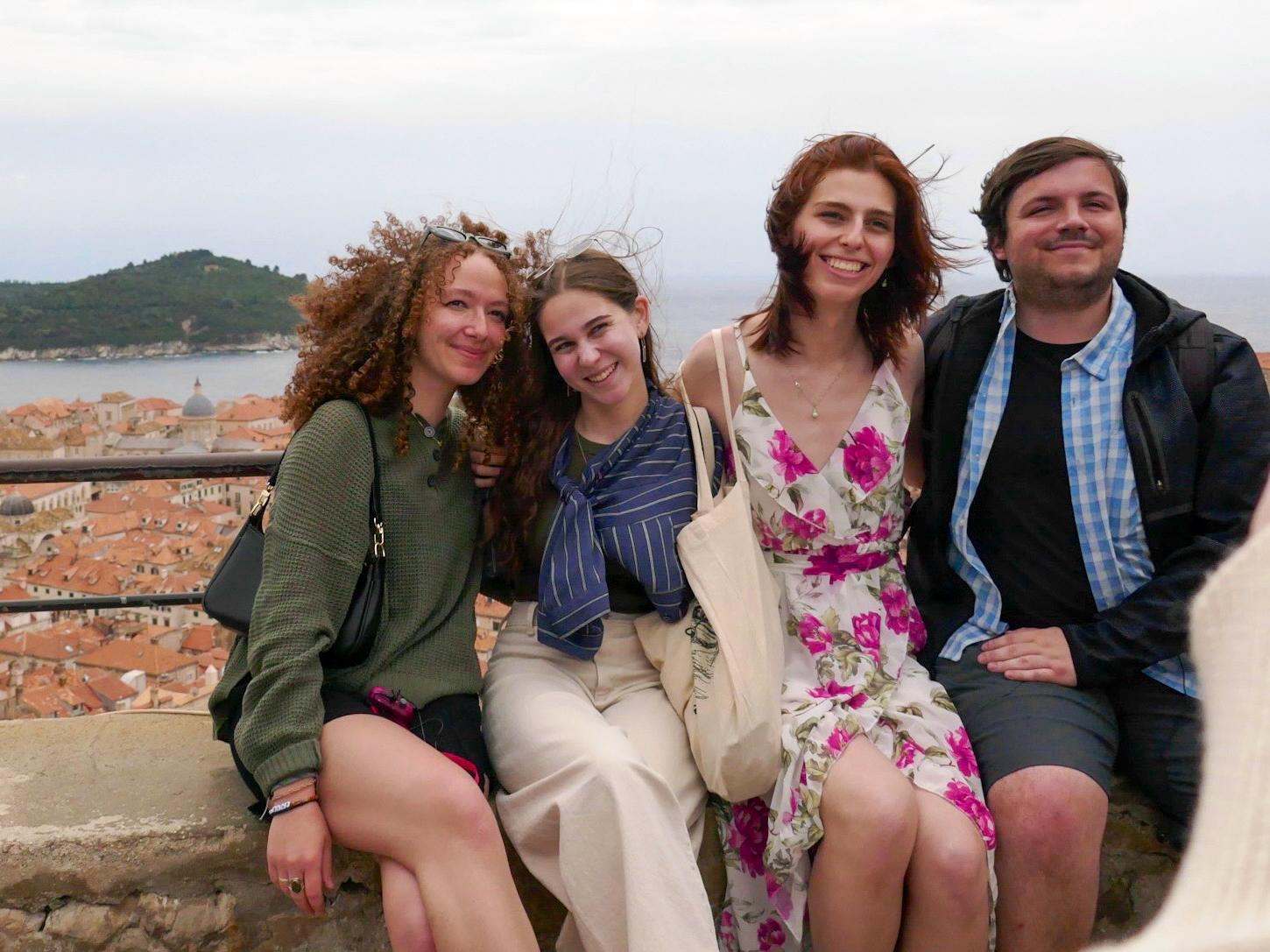 Three young women and one young man sit on a concrete ledge overlooking  Dubrovnik, Croatia, dressed in spring clothing.