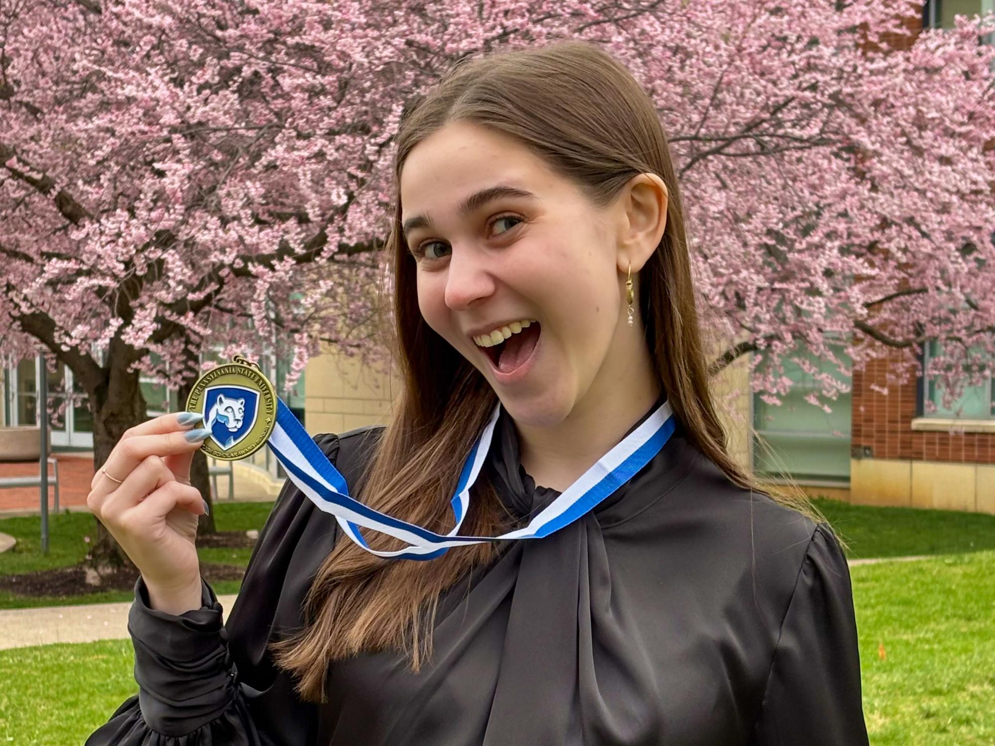 Emily Danczyk stands outdoors in front of blooming pink cherry blossom trees, smiling and holding a medal on a blue-and-white ribbon. 