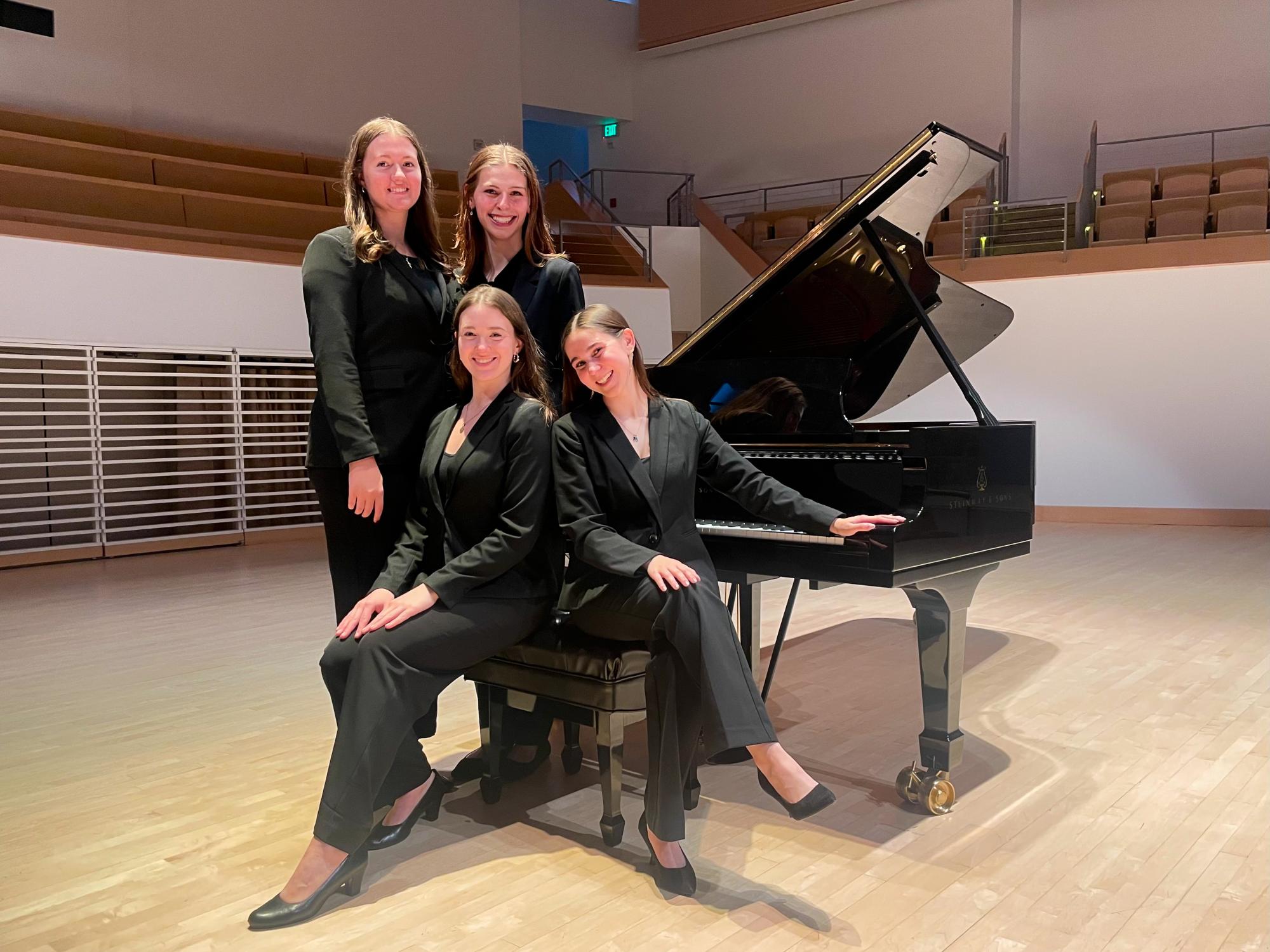 Four young women dressed in black formal attire pose beside a grand piano on a recital hall stage.