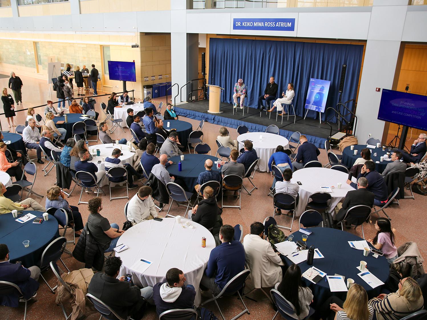 A view of a crowd in the Business Building atrium listening to three speakers on a stage.