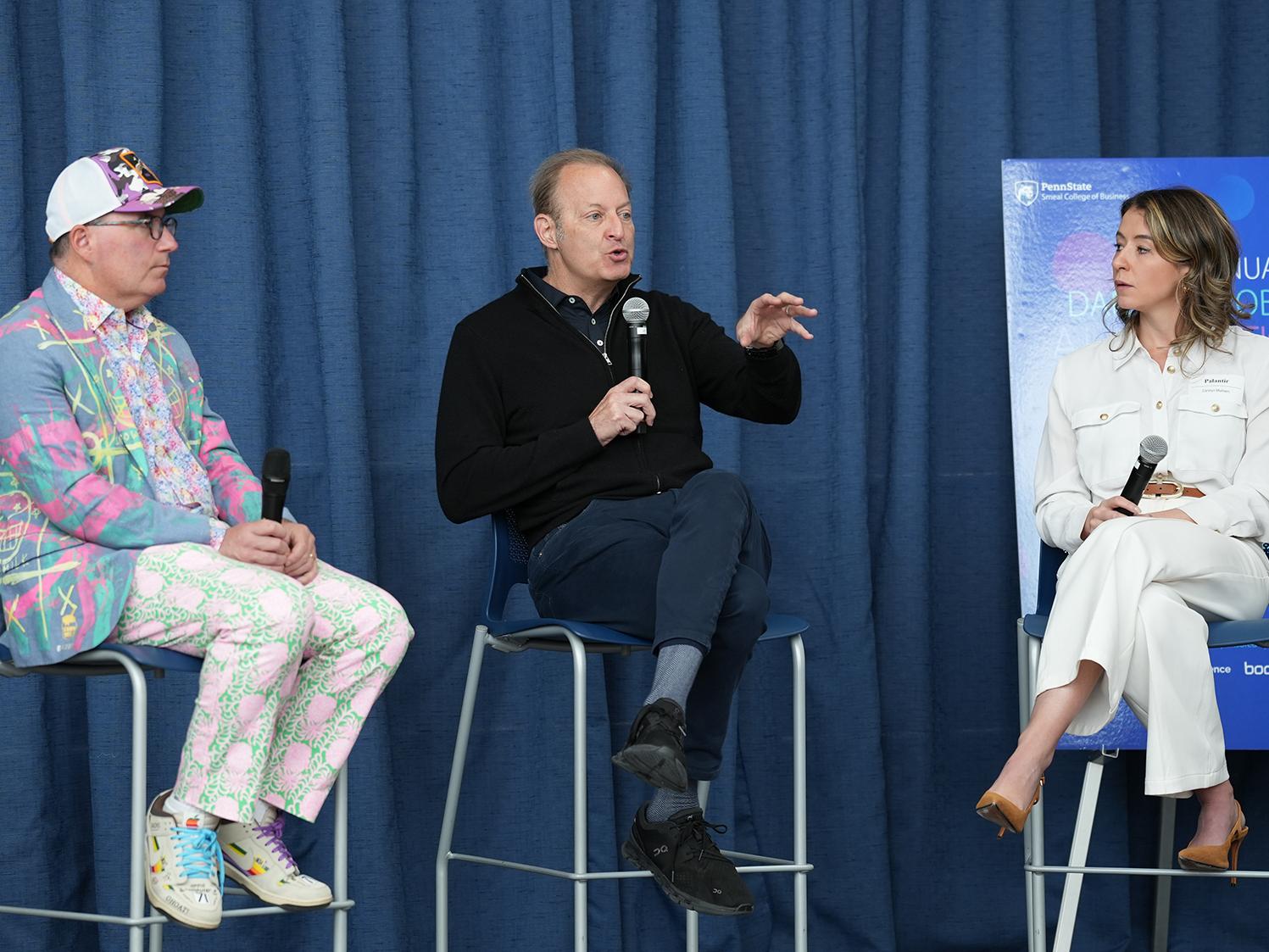 Two men, Dan Ives and David Steinberg, sit on stools with a woman, Carolyn Mulhern, on a stage talking.