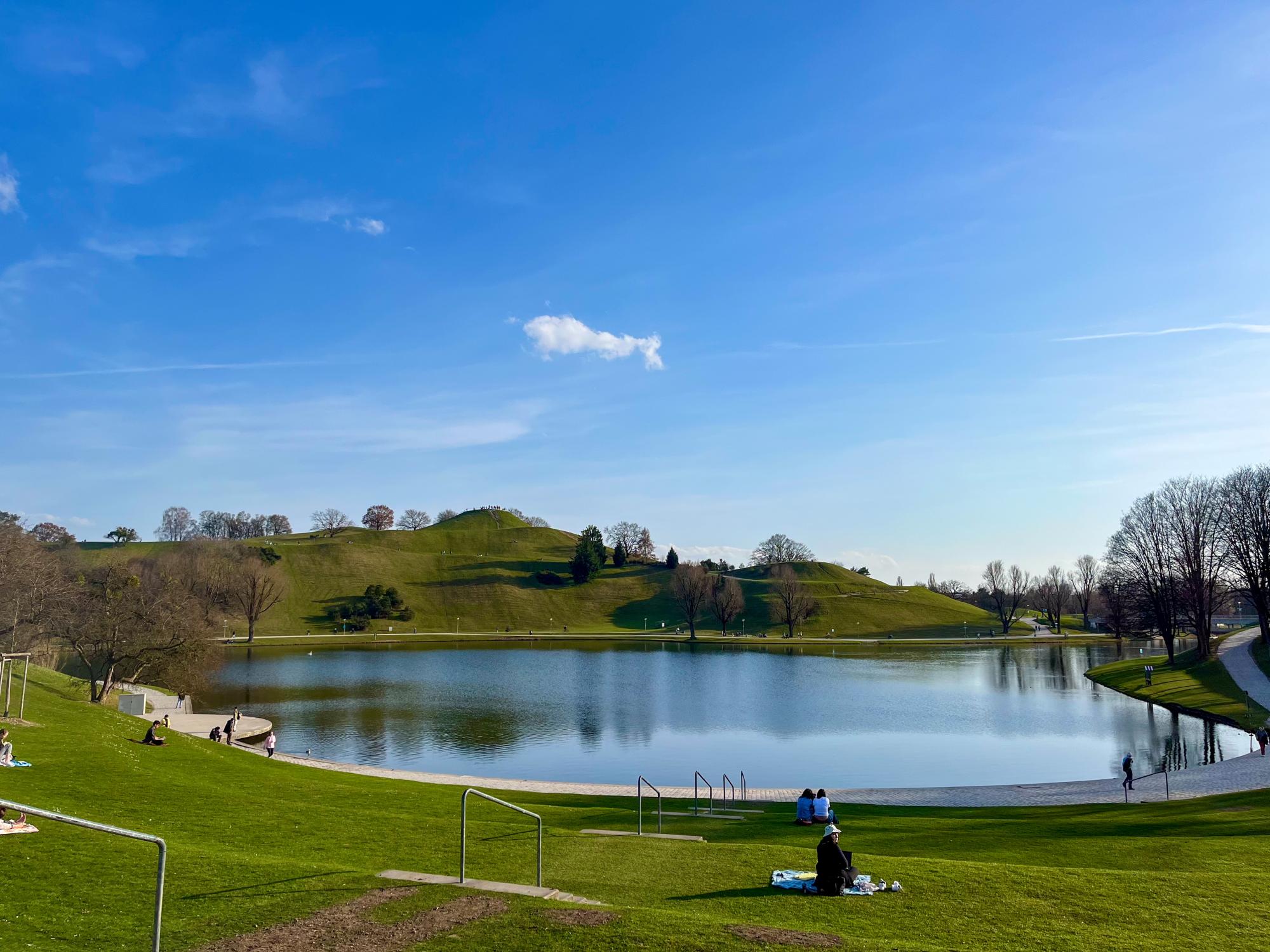 A landscape photo of the Olympic Park and its lake, Olympiasee, taken outside on a sunny day. 