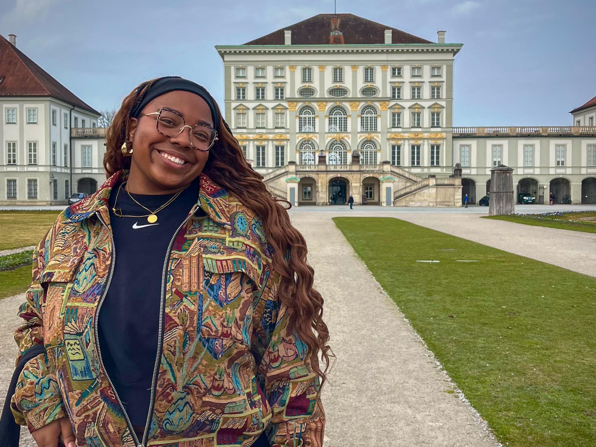 A young woman in a colorful zip-up poses in front of a Baroque palace, smiling on a cloudy day.