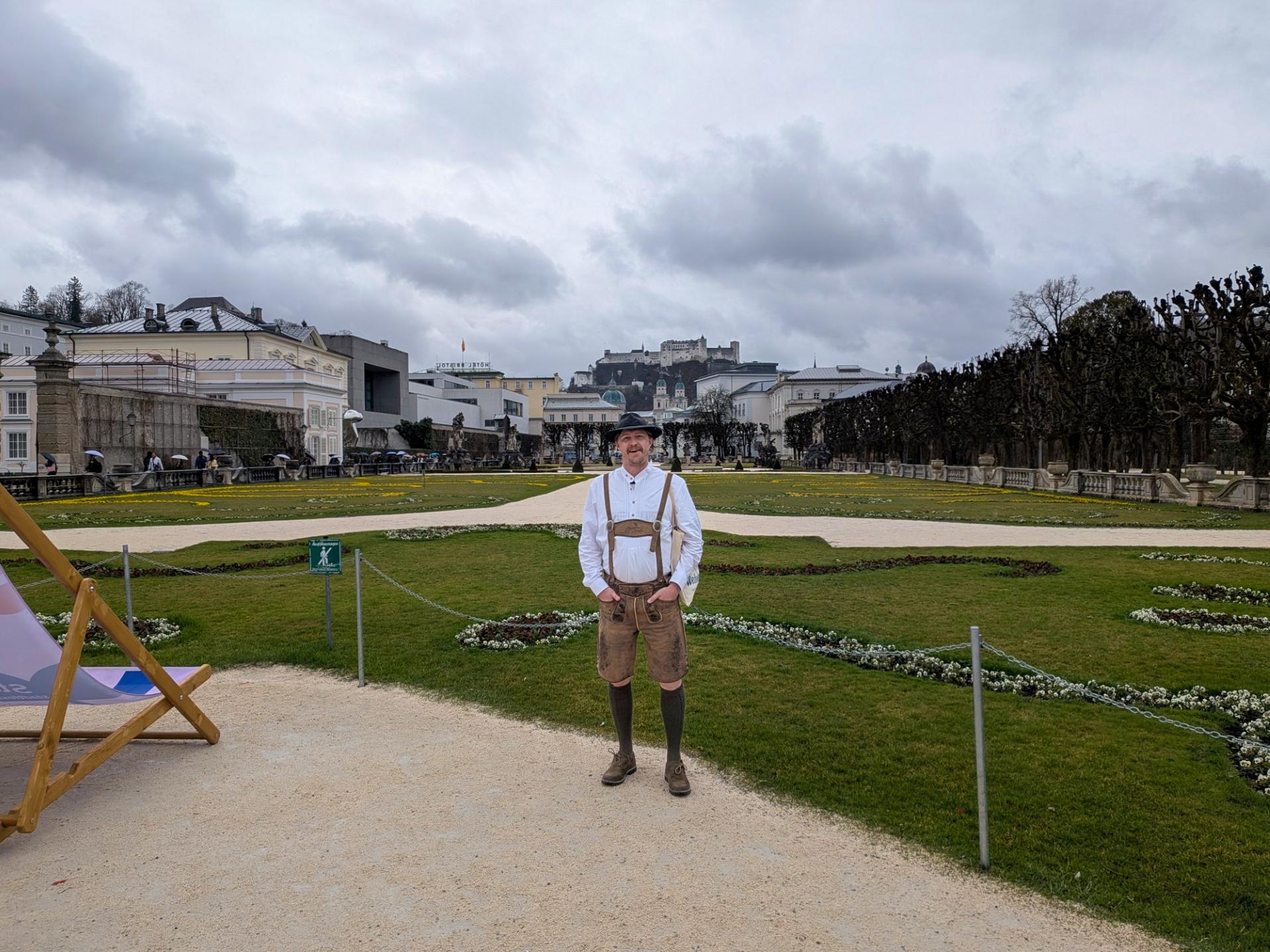 A man stands outside, hands in his pockets, in front of a green landscape, wearing a white shirt, brown lederhosen, and a felt fedora.