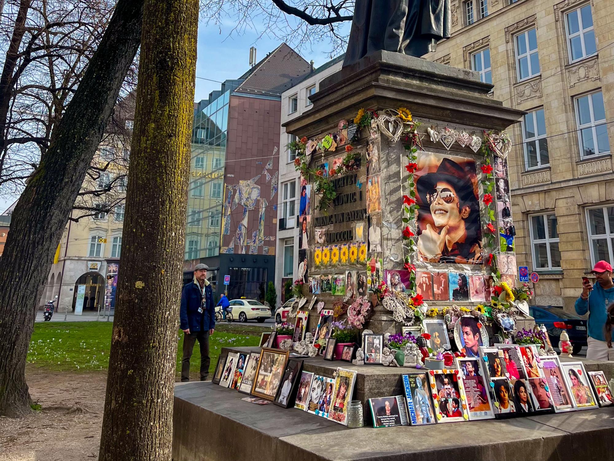 A collection of flowers and Michael Jackson pictures placed around the base of a statue on a sunny day.