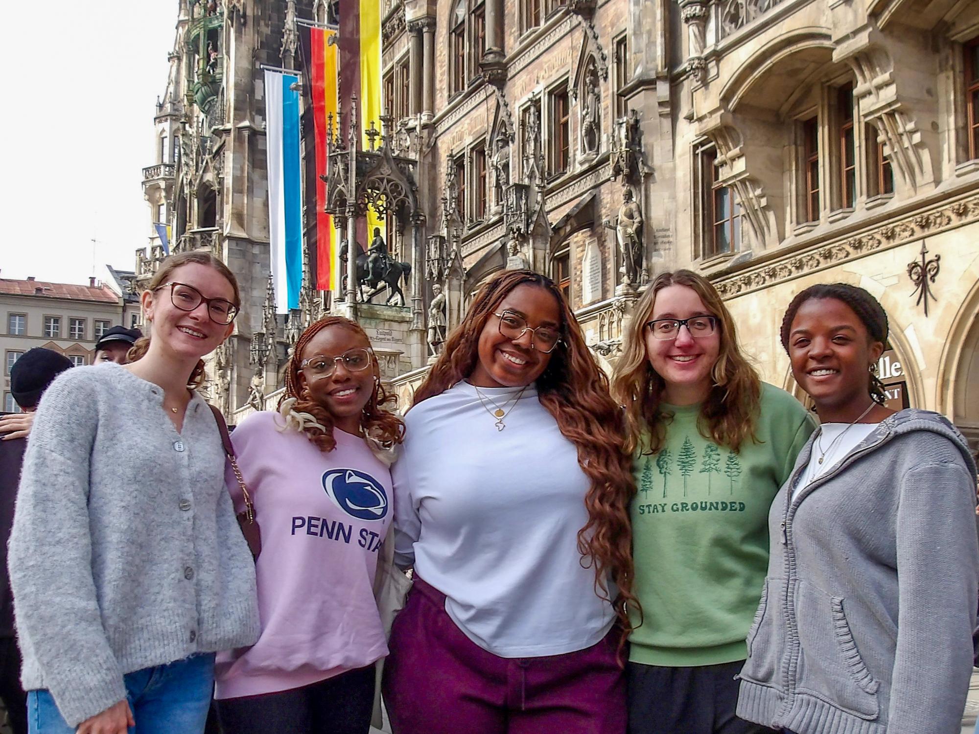 Five young women dressed in loungewear pose in front of the Munich Town Hall on a sunny day.