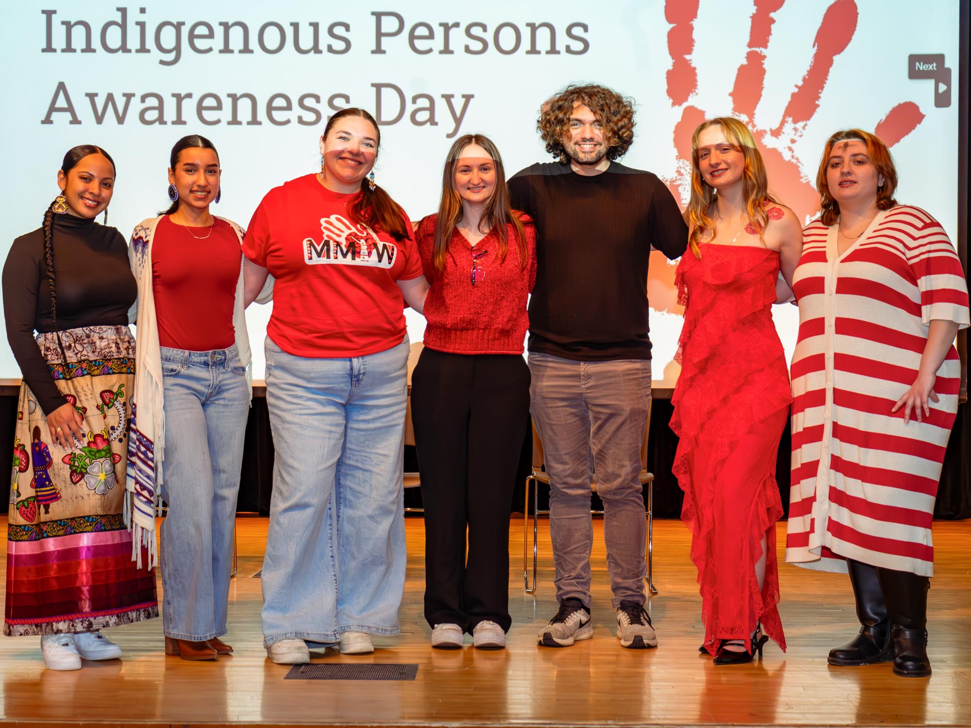 Laenee McCoy (second to last on the right) and other IPSA members stand in front of the Indigenous Persons Awareness Day sign. Laenee is wearing a long red dress.