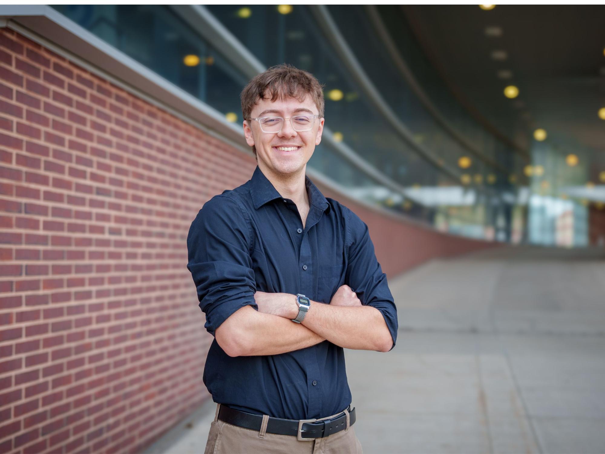 person in black shirt and khakis stands with arms crossed near brick wall